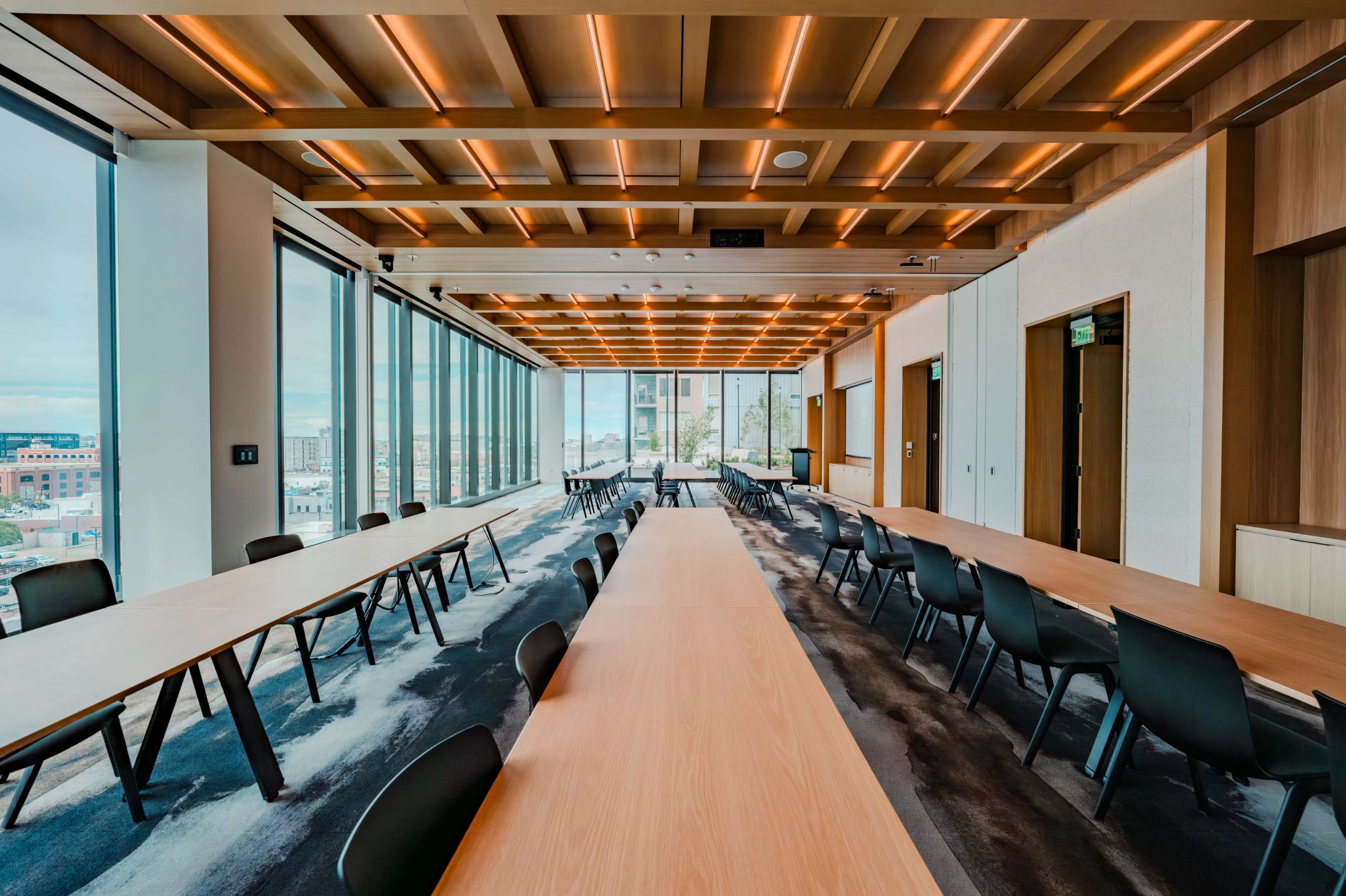 A modern conference room features large windows, wooden beams on the ceiling, and multiple long tables arranged with black chairs.