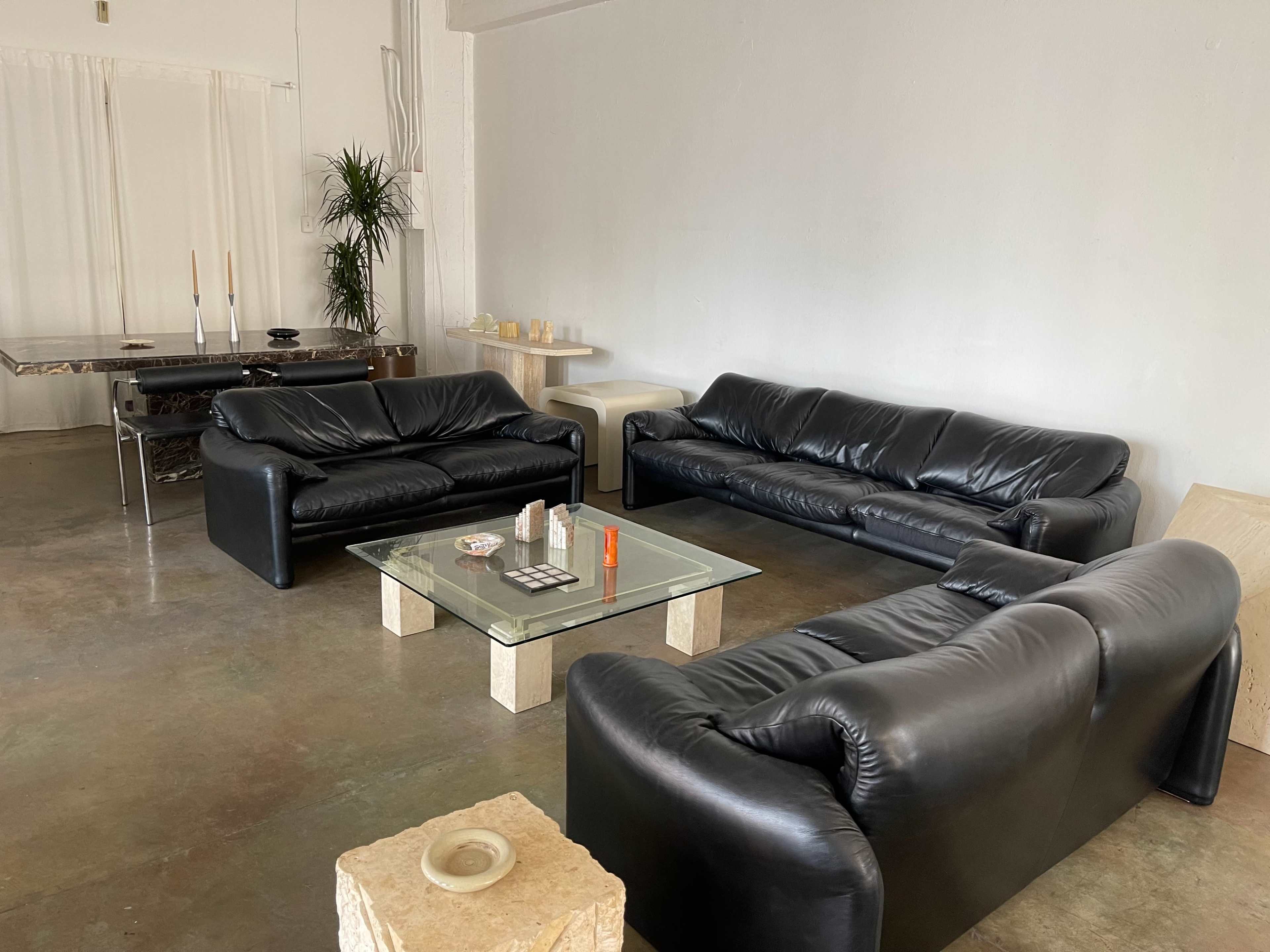 A modern interior space featuring three black leather sofas arranged around a glass coffee table on a concrete floor.