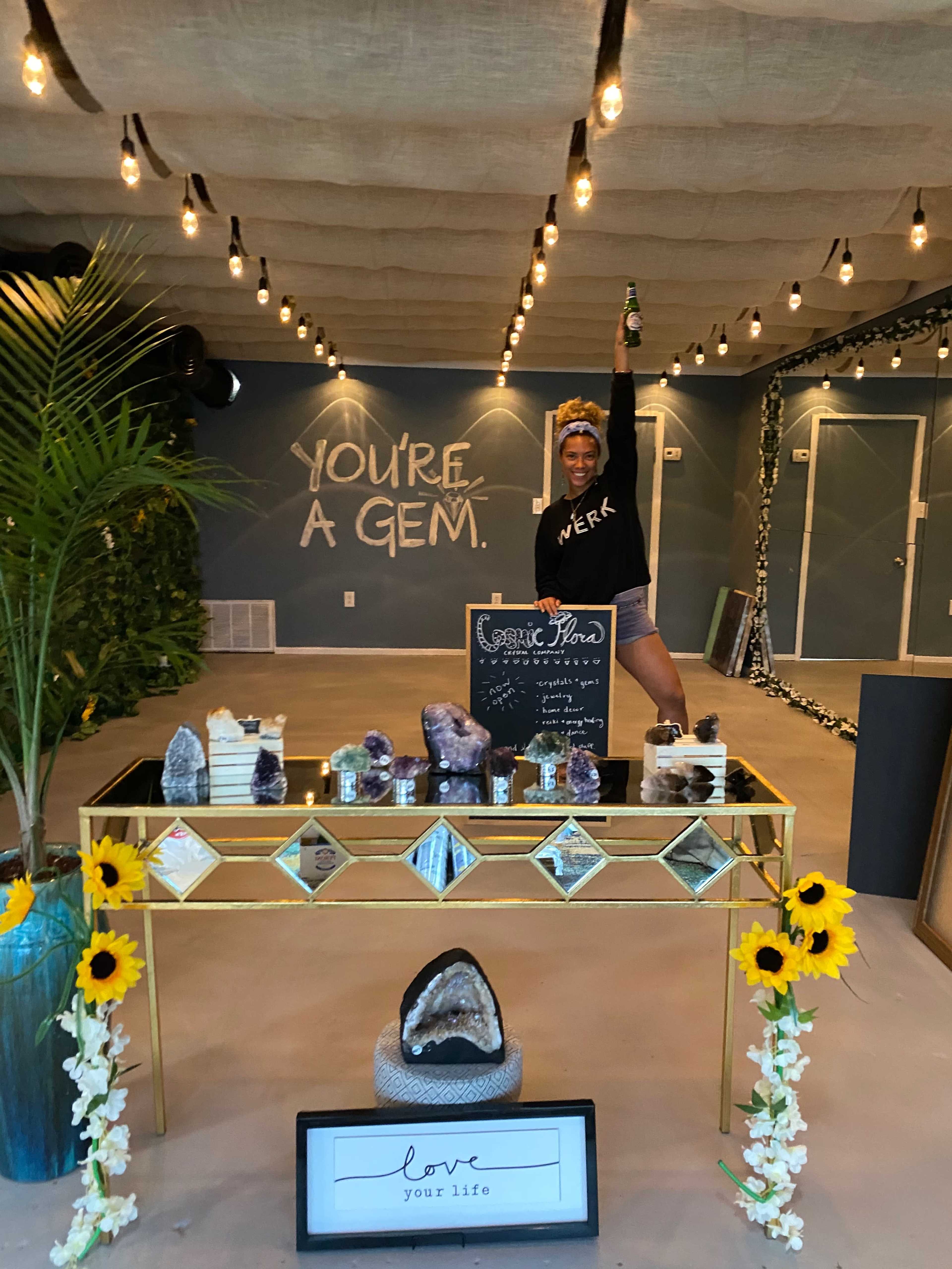 A person stands with one arm raised behind a table displaying various crystals and decorative items in a brightly lit space.