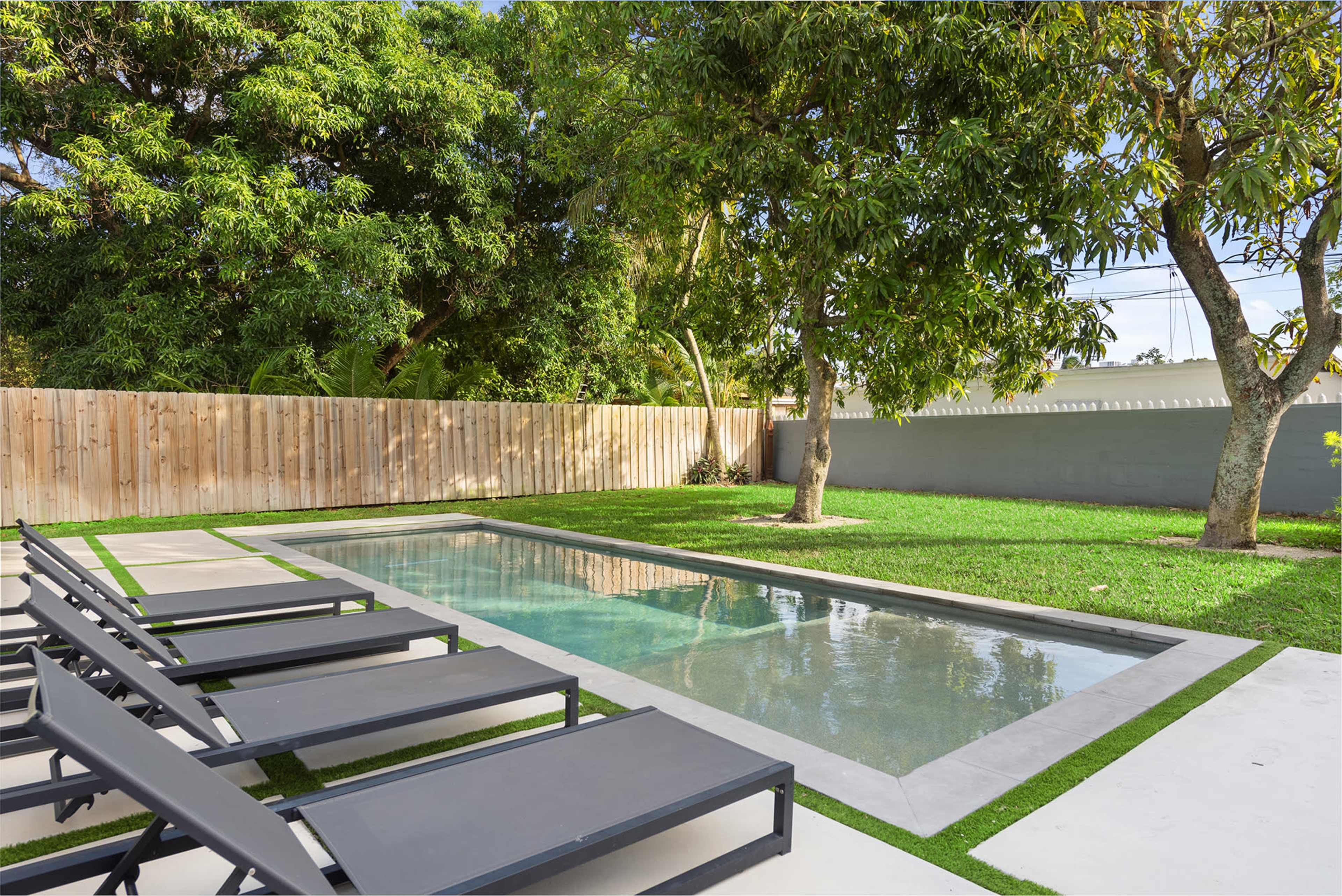 The image shows a modern backyard with a rectangular swimming pool surrounded by lounge chairs and grass, enclosed by a wooden fence and trees.