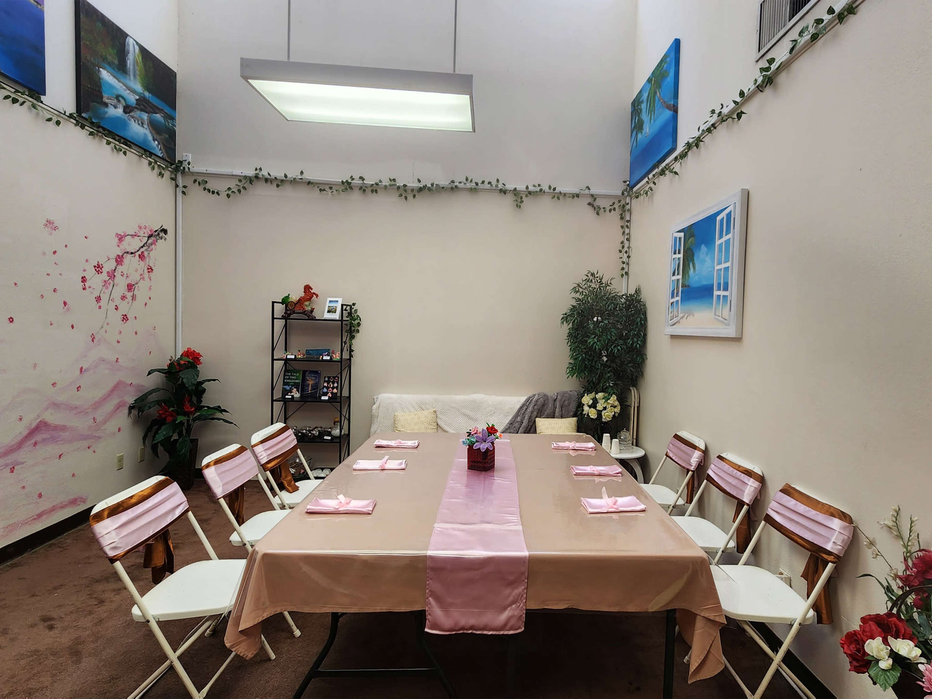 The image shows a decorated room with a long table set for a meal, surrounded by white folding chairs and adorned with potted plants and wall art.