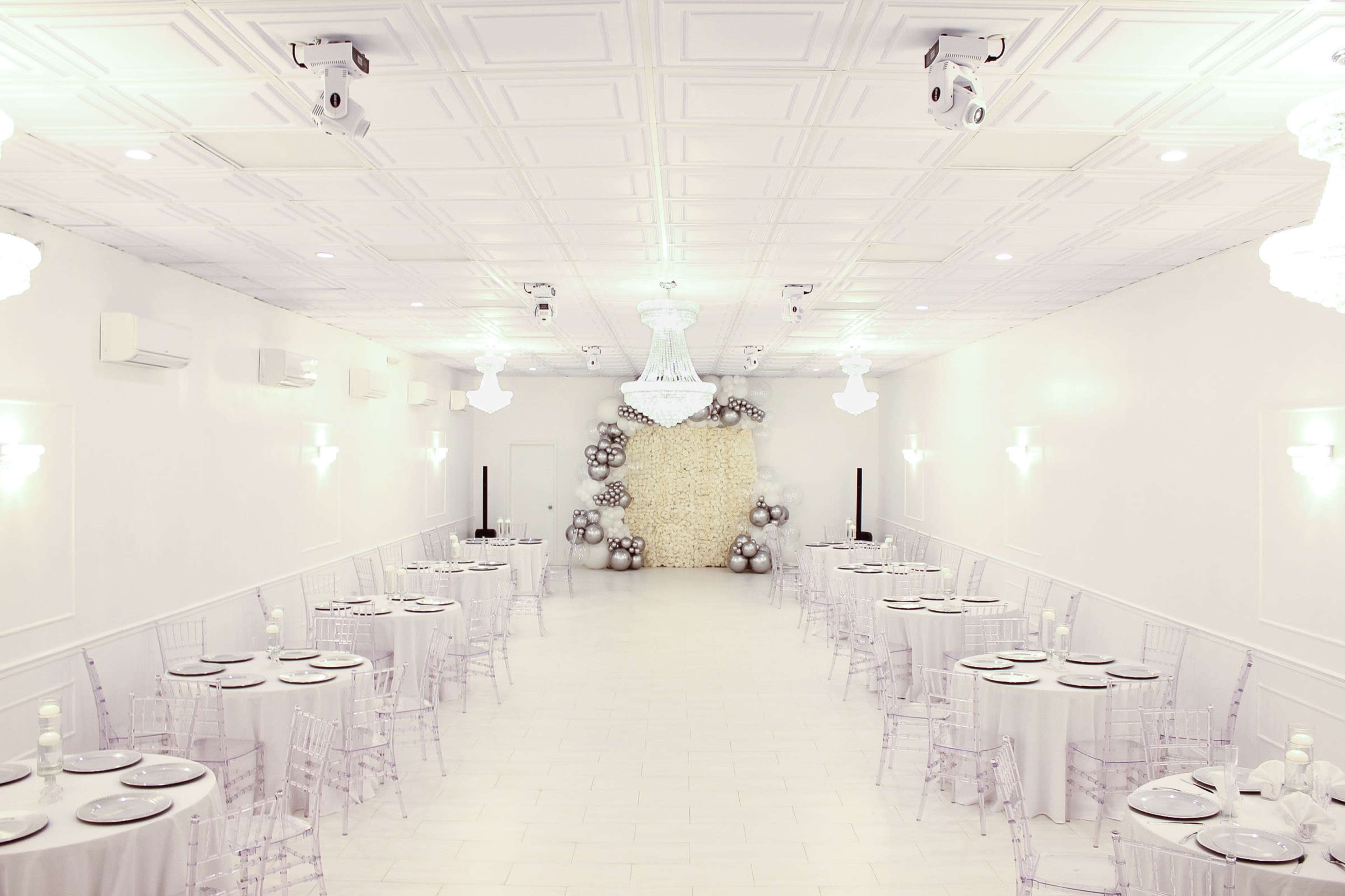 A spacious, elegantly set dining hall with round tables covered in light-colored tablecloths, transparent chairs, and a decorative wall featuring a cluster of balloons.