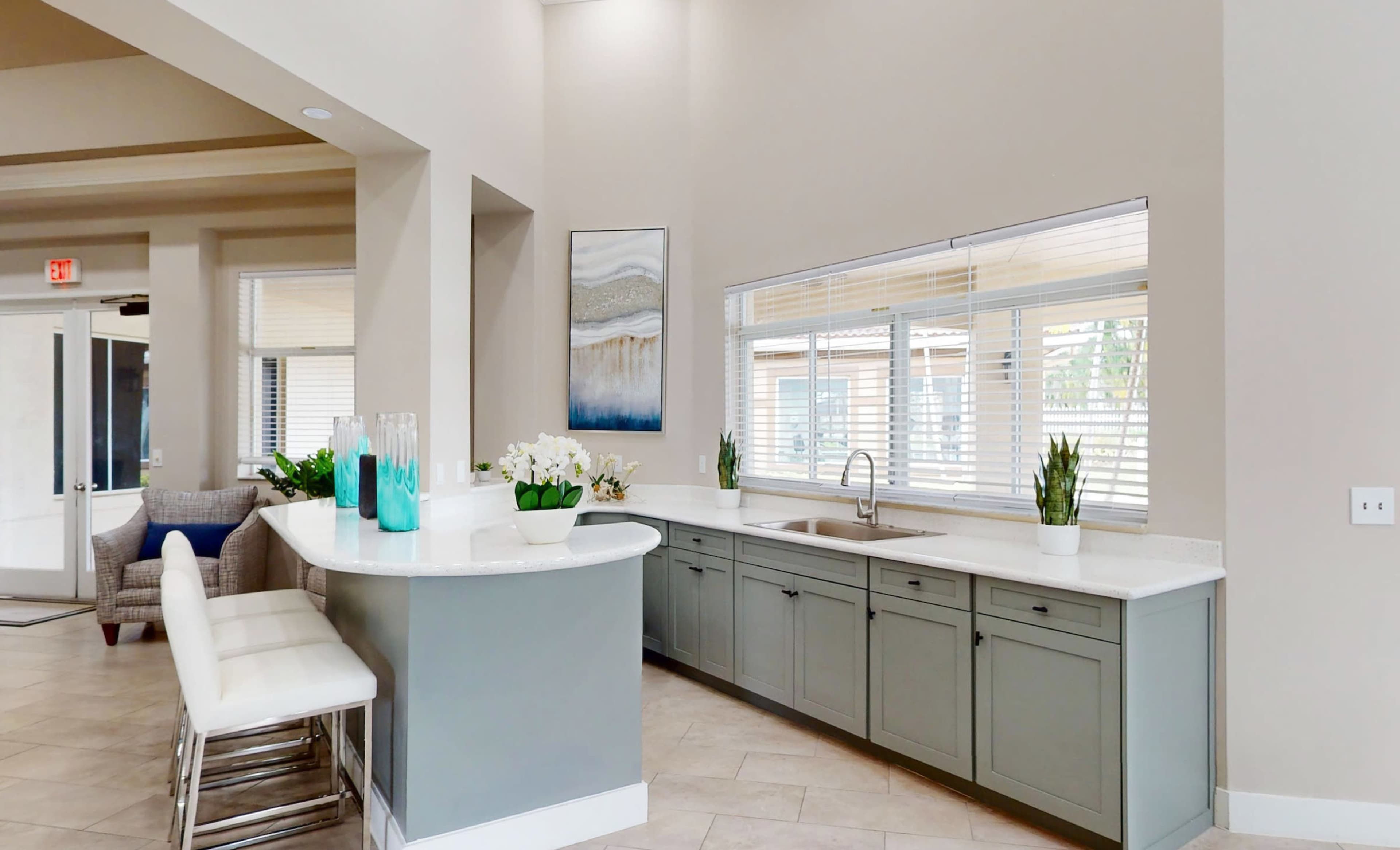 The image shows a modern kitchen with a gray island, white countertop, and large windows allowing natural light to fill the space.