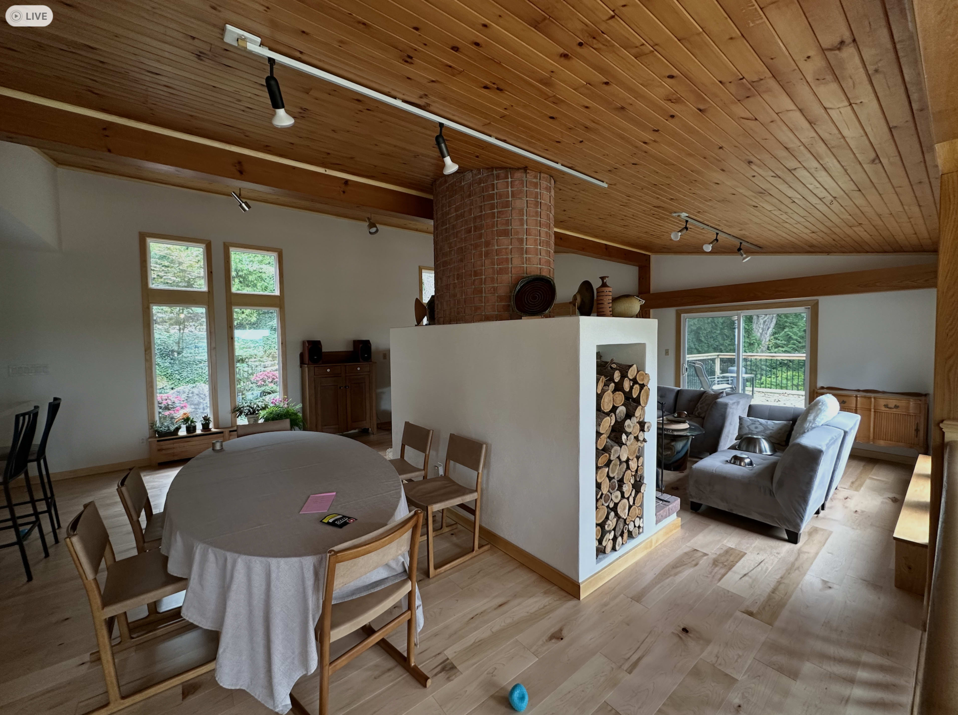 The image shows a modern, open-concept living space with a dining area featuring a round table, a seating area with a gray couch, and a central fireplace stacked with logs.