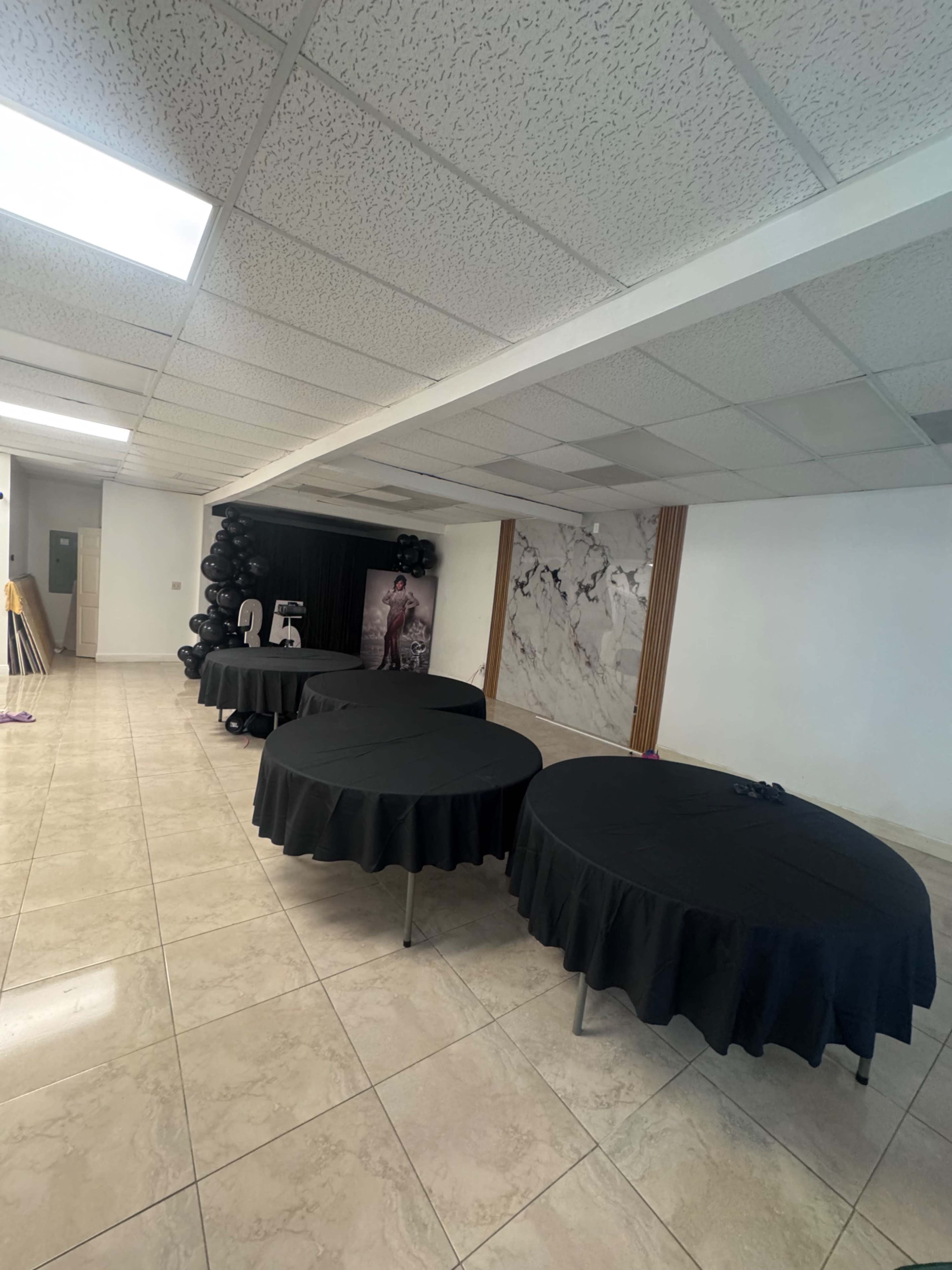 The image shows a spacious indoor venue with several round tables covered in black tablecloths, along with a wall featuring marble accents and black balloons.