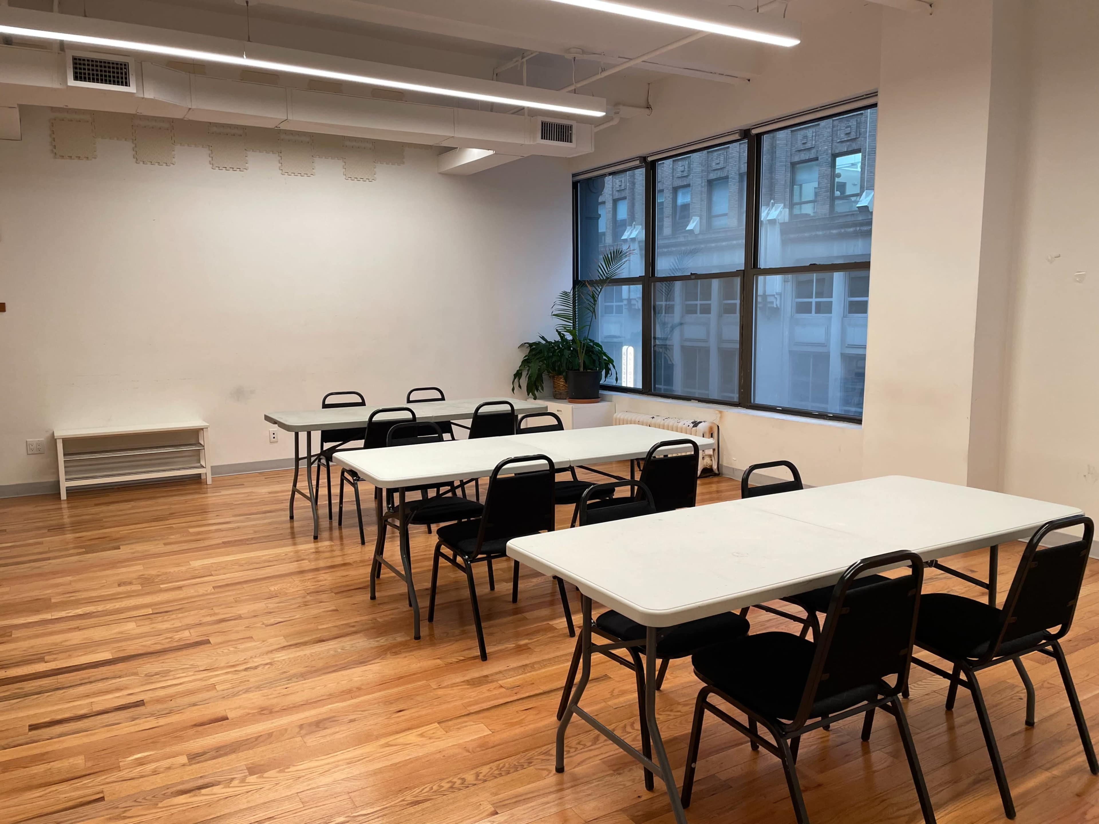 A bright room features multiple black chairs arranged around white tables on wooden flooring, with large windows and a potted plant in the corner.