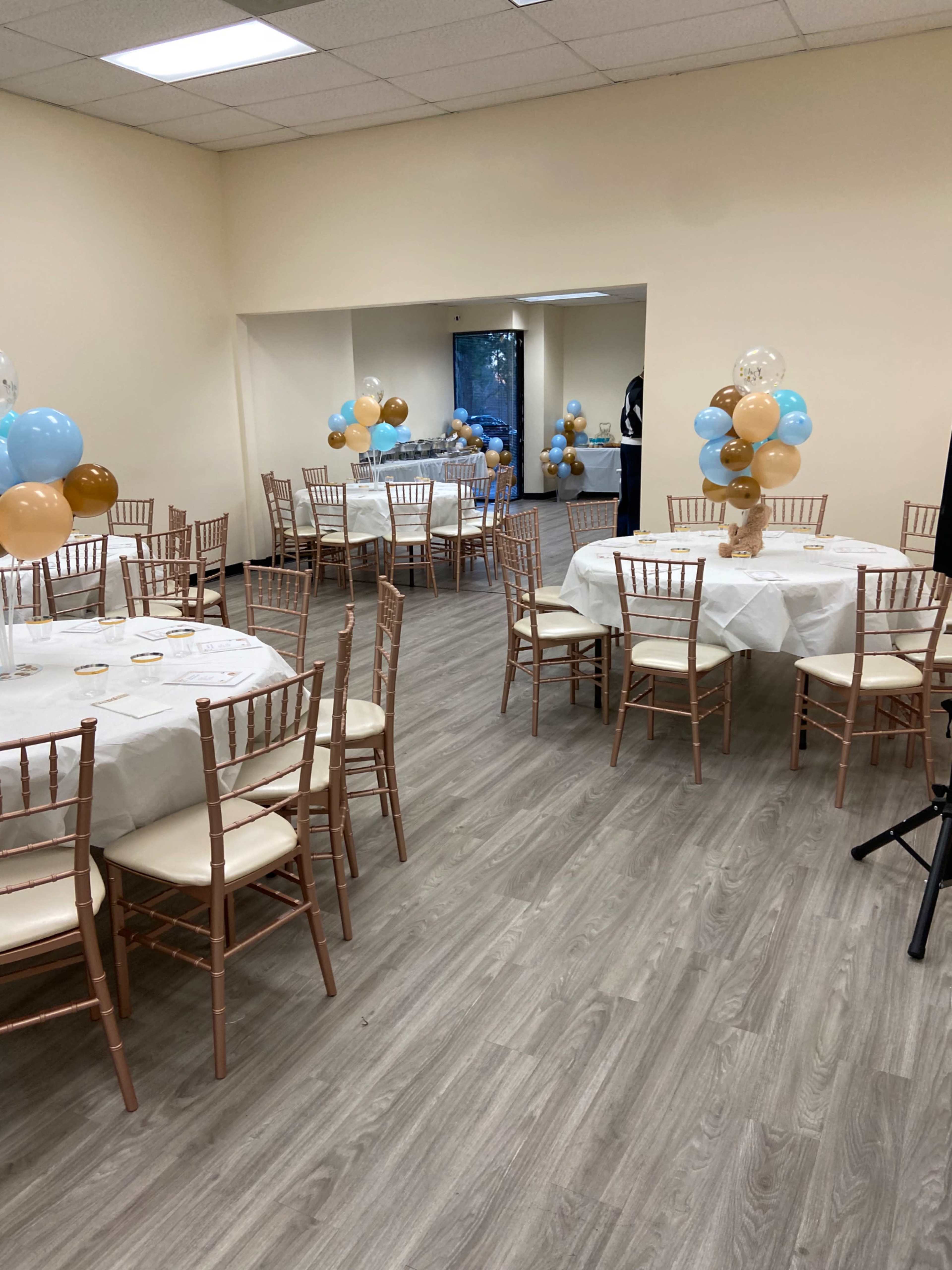 The image shows a hall with several round tables covered in white tablecloths, each surrounded by wooden chairs, and decorated with balloons in blue, brown, and clear colors.