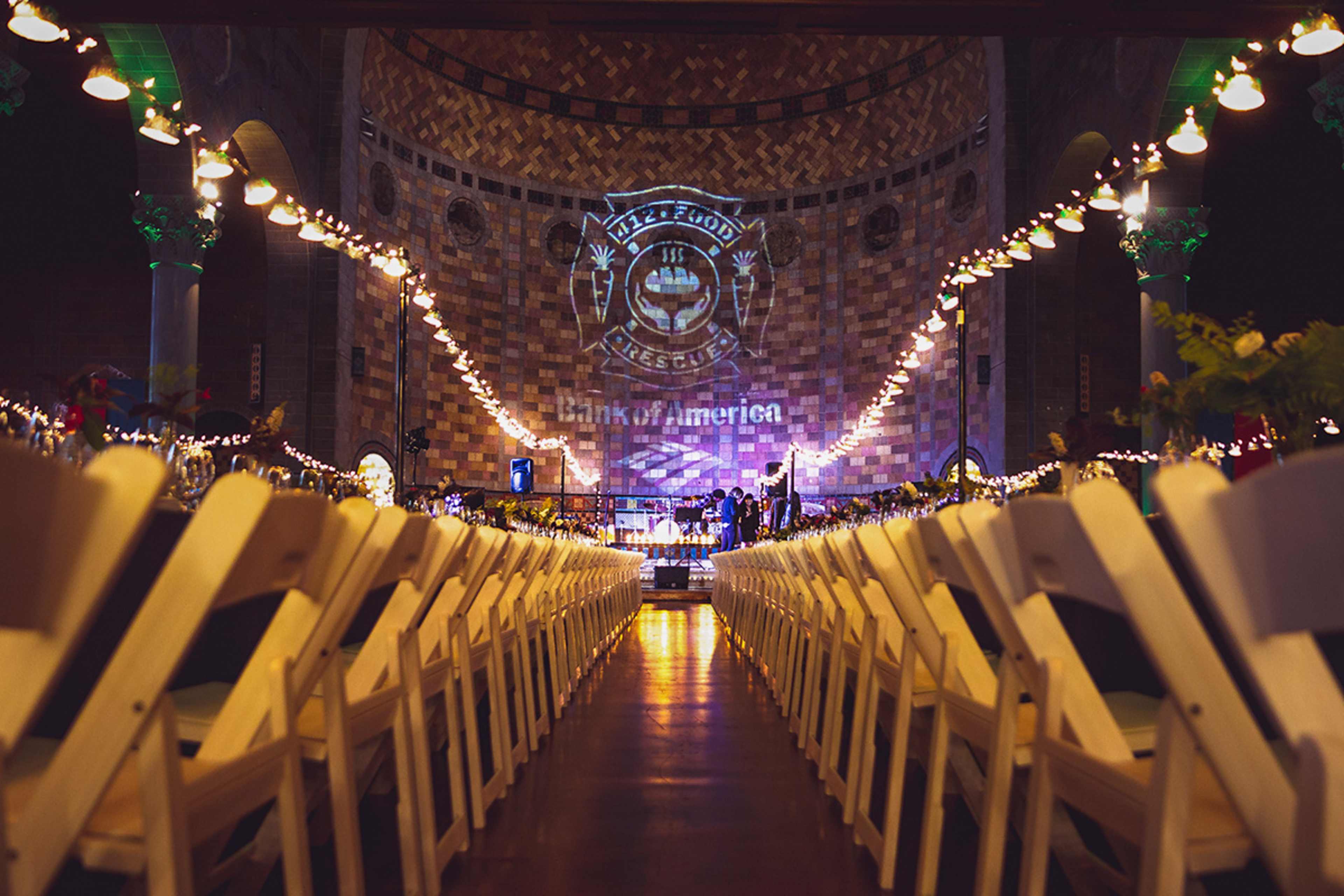An interior event space features rows of white chairs set up for a gathering, with a large projection on the wall displaying a logo and the words "Bank of America."