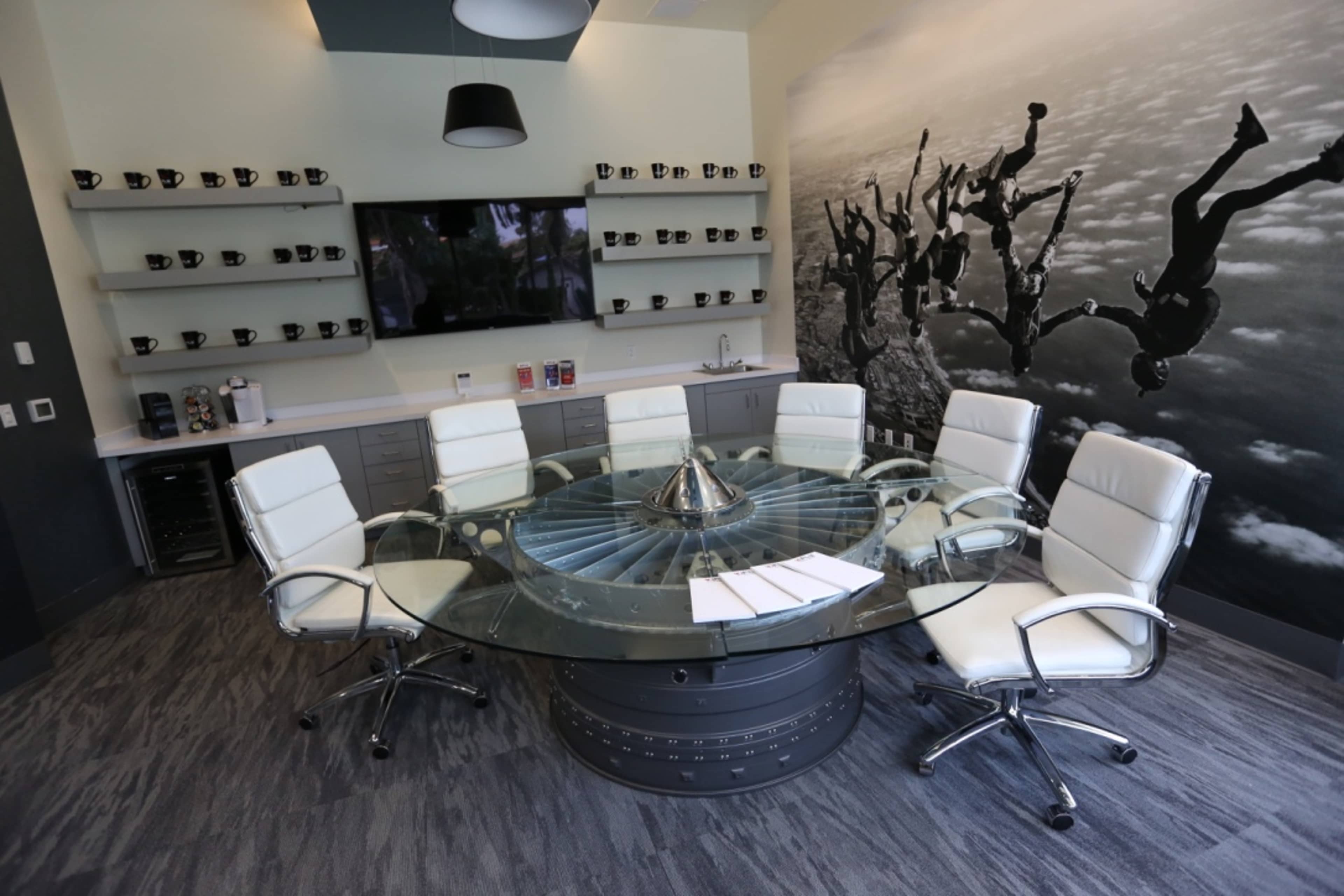 A modern conference room features a glass circular table surrounded by white leather chairs, with a wall mural of people skydiving and shelves displaying coffee mugs.