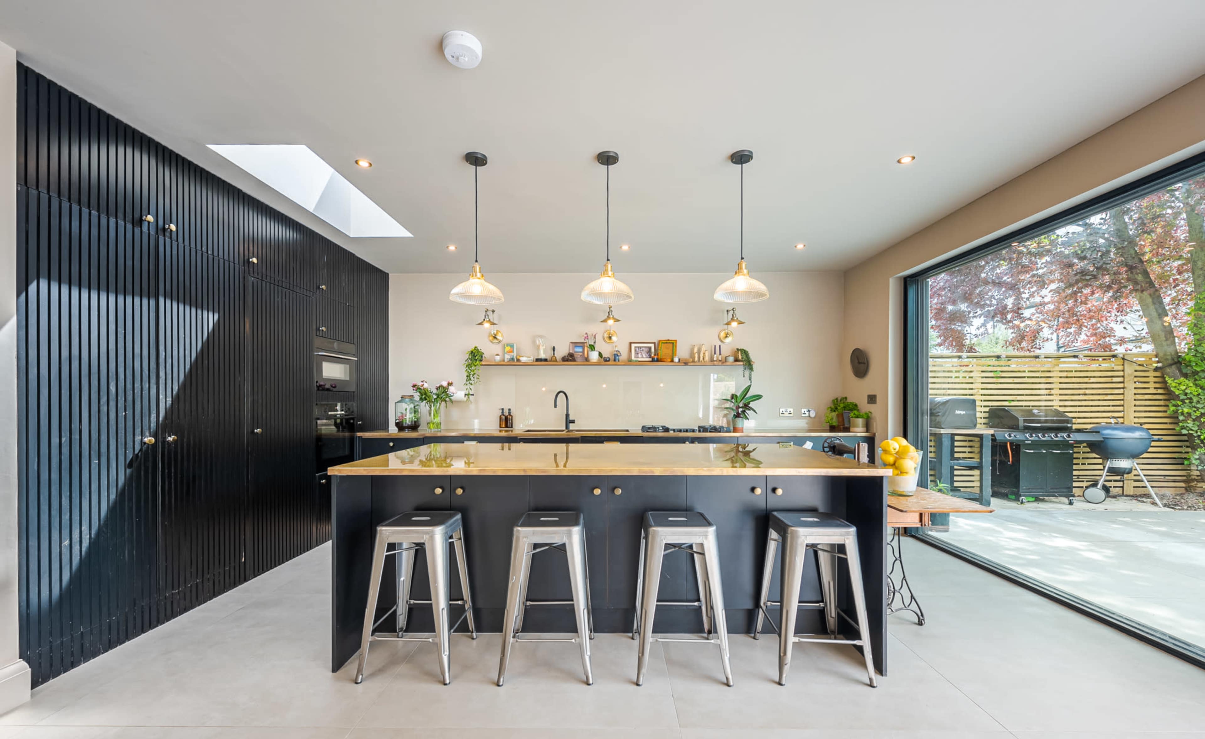 The image shows a modern kitchen featuring a central island with four metal stools, a sleek countertop, and large windows that provide a view of an outdoor area.