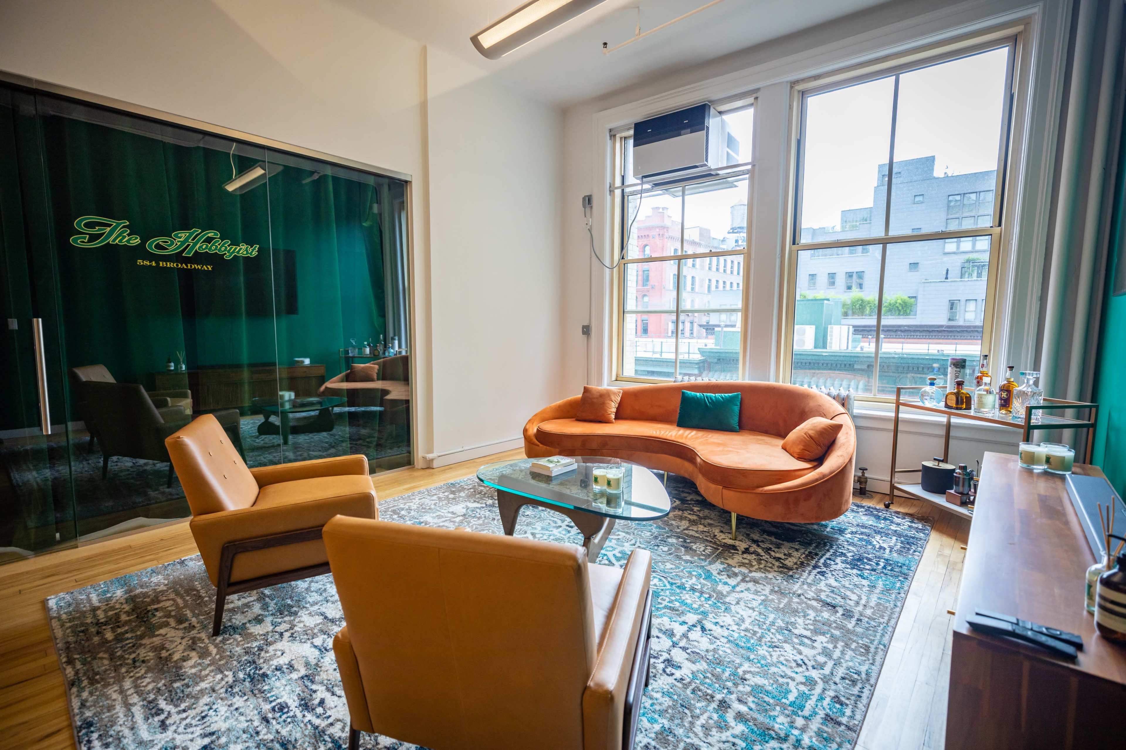 A modern lounge area with a curved orange sofa, two brown chairs, a patterned rug, and large windows overlooking a cityscape.