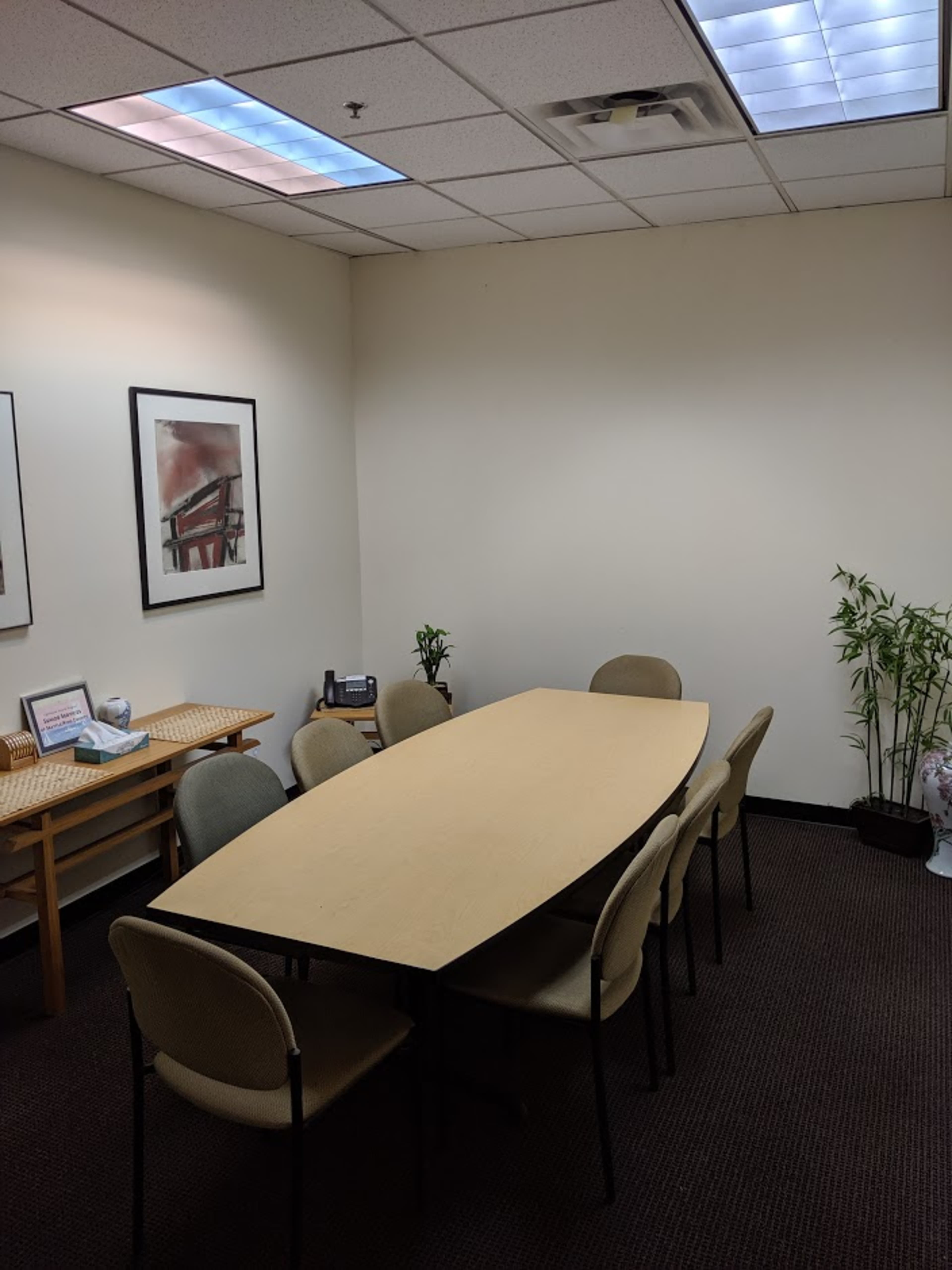 A conference room features a long table surrounded by chairs, with framed artwork on the walls and plants in the corners.