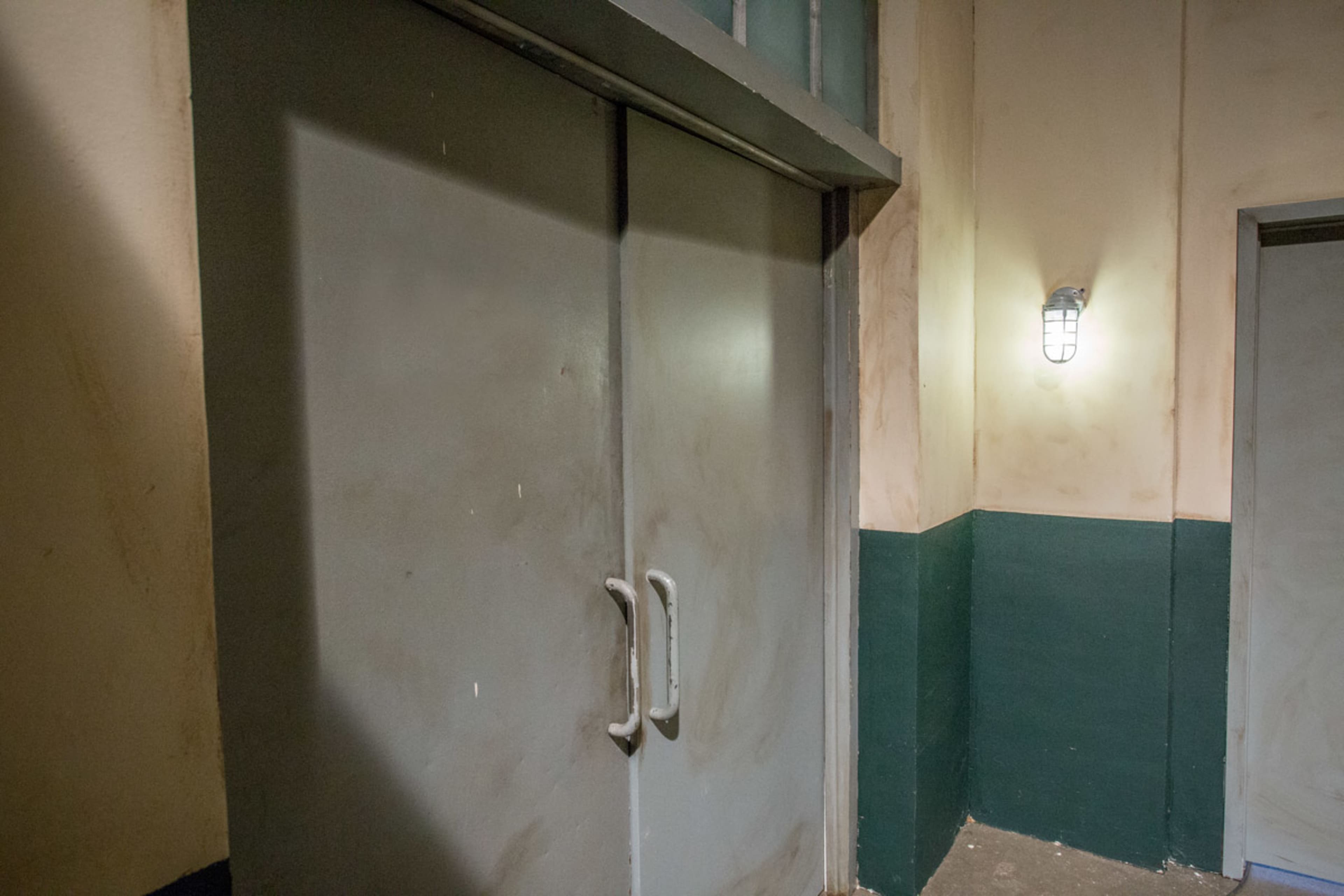 A pair of gray double doors is positioned in a dimly lit corridor with textured beige walls and a green lower section.