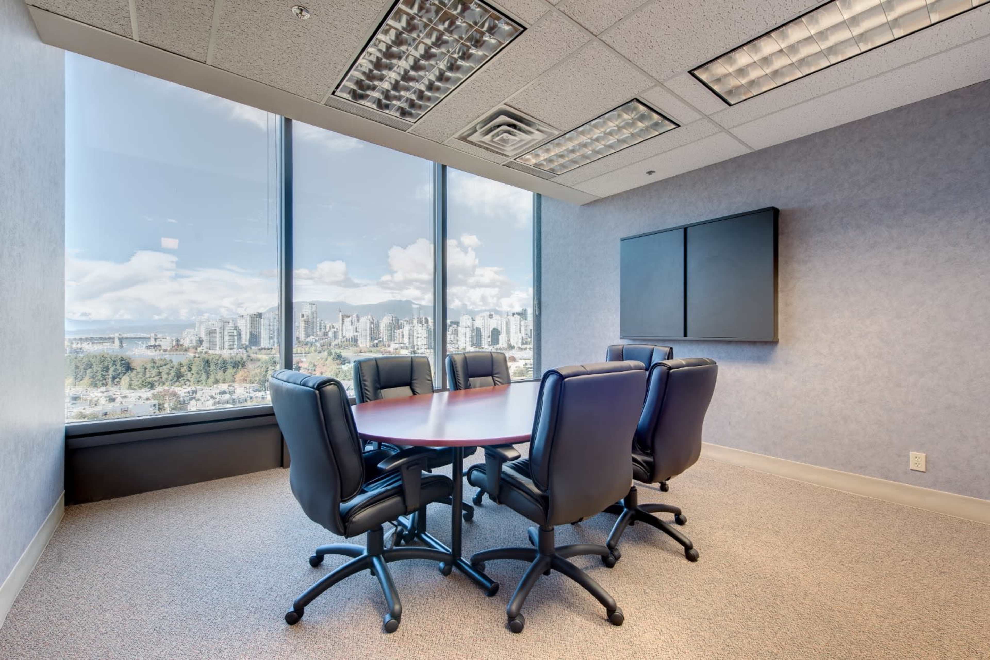 A conference room with a round table surrounded by six black office chairs, featuring large windows that overlook a city skyline.