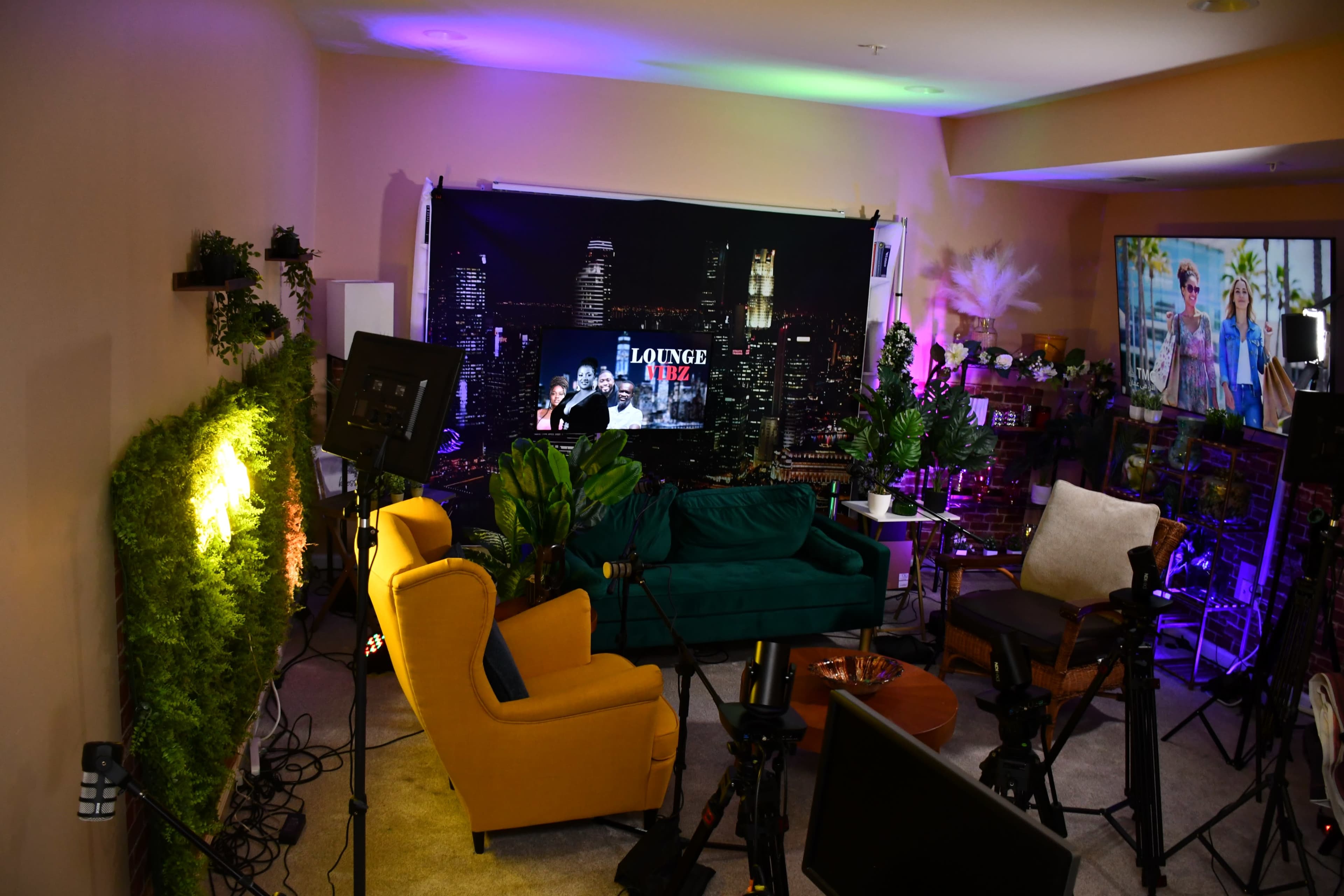 The image shows a brightly lit lounge area equipped with various seating, plants, and filming equipment, set against a backdrop featuring a city skyline.