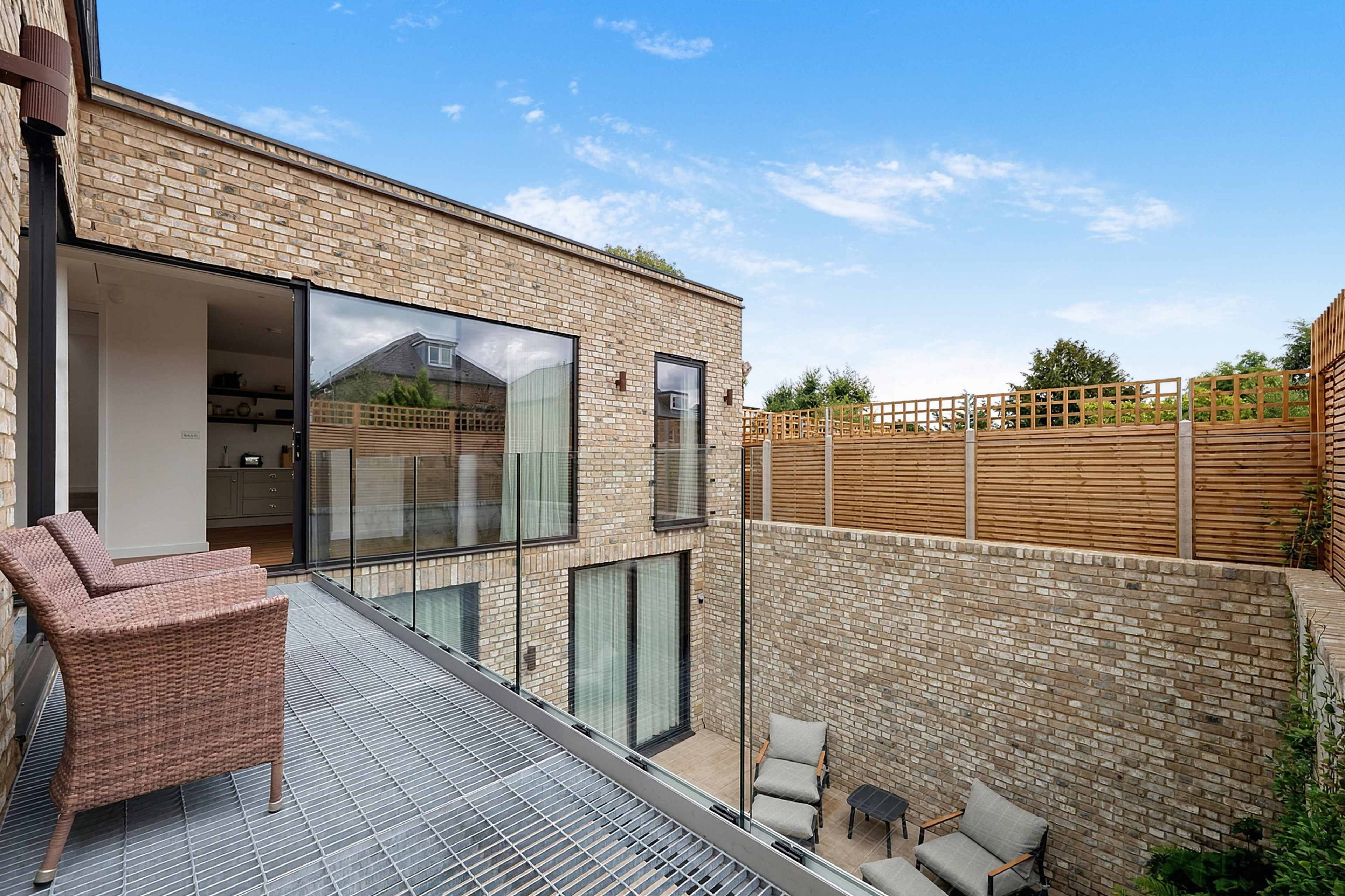 The image shows a modern courtyard with a brick exterior, featuring a balcony with two chairs, and a fenced area with seating and a small table.