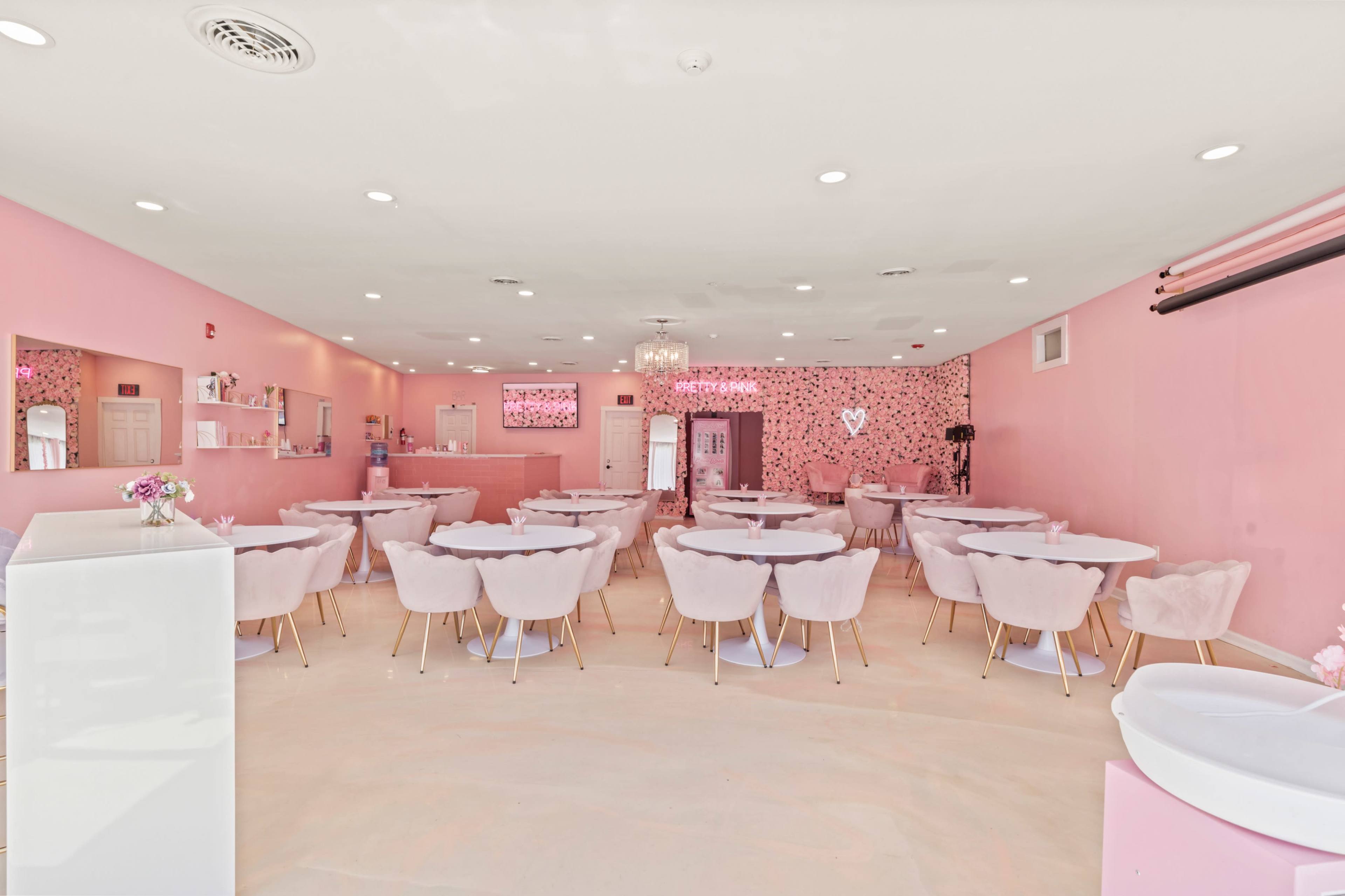 A pink-themed café with round tables and chairs, floral wall decor, and a bar area at the back.