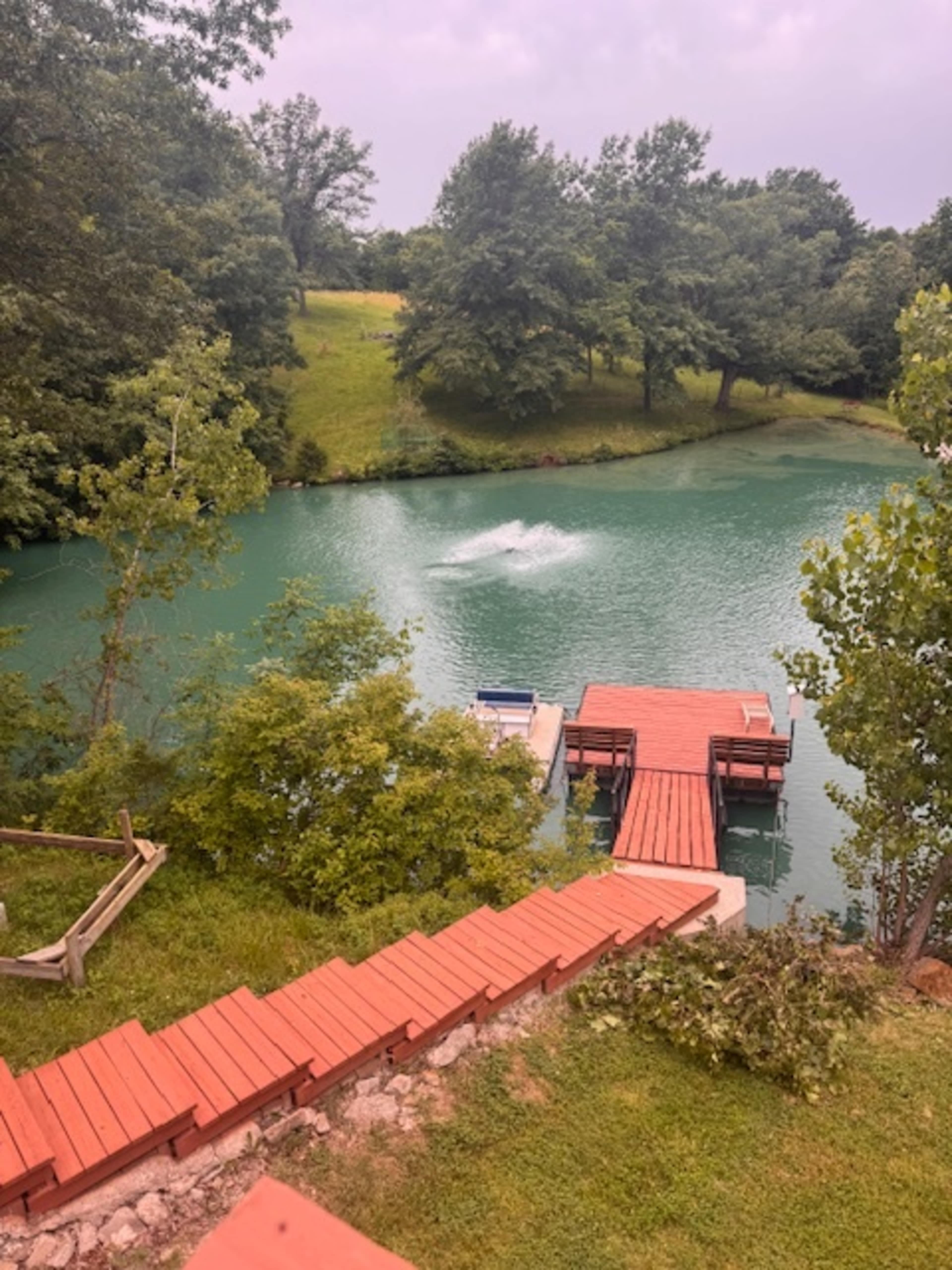 The image shows a staircase leading down to a wooden dock on a turquoise pond surrounded by green trees.