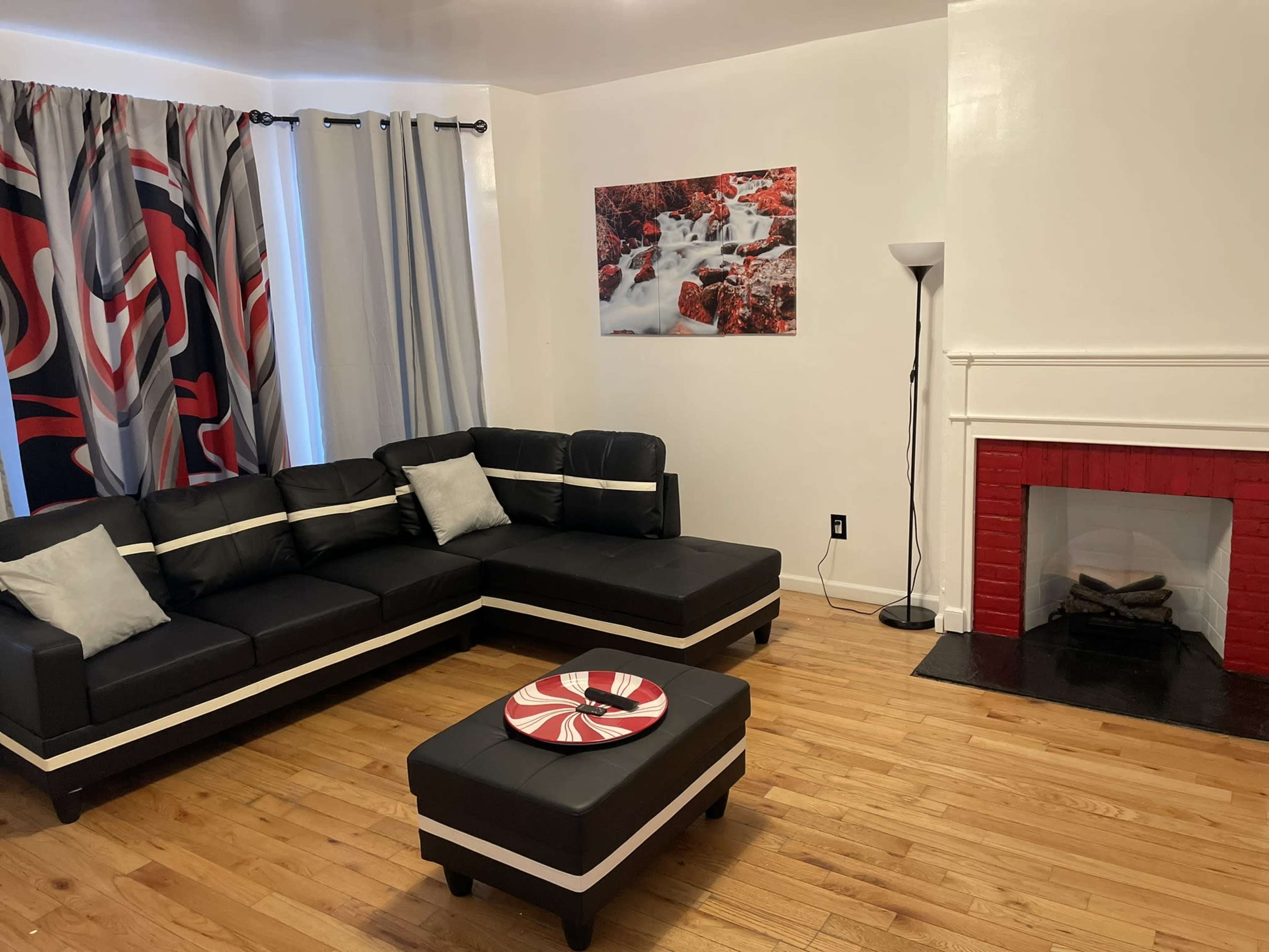 The living room features a black sectional couch, a round coffee table with a decorative plate, a red fireplace, and a large photograph of a water scene on the wall, with curtains displaying a bold pattern.