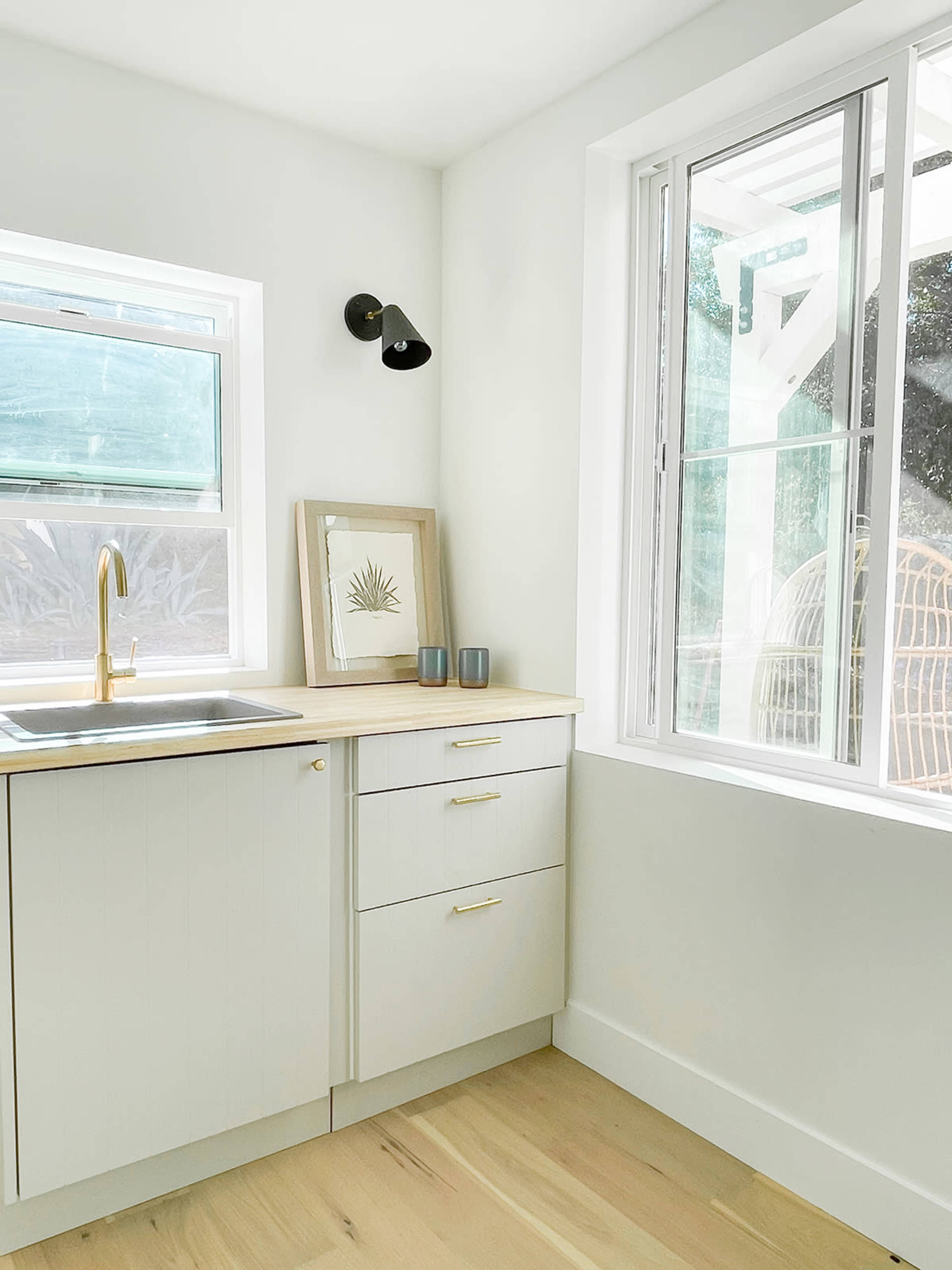 The image shows a modern kitchen corner with a sink, wooden countertop, framed artwork, and natural light coming through a window.