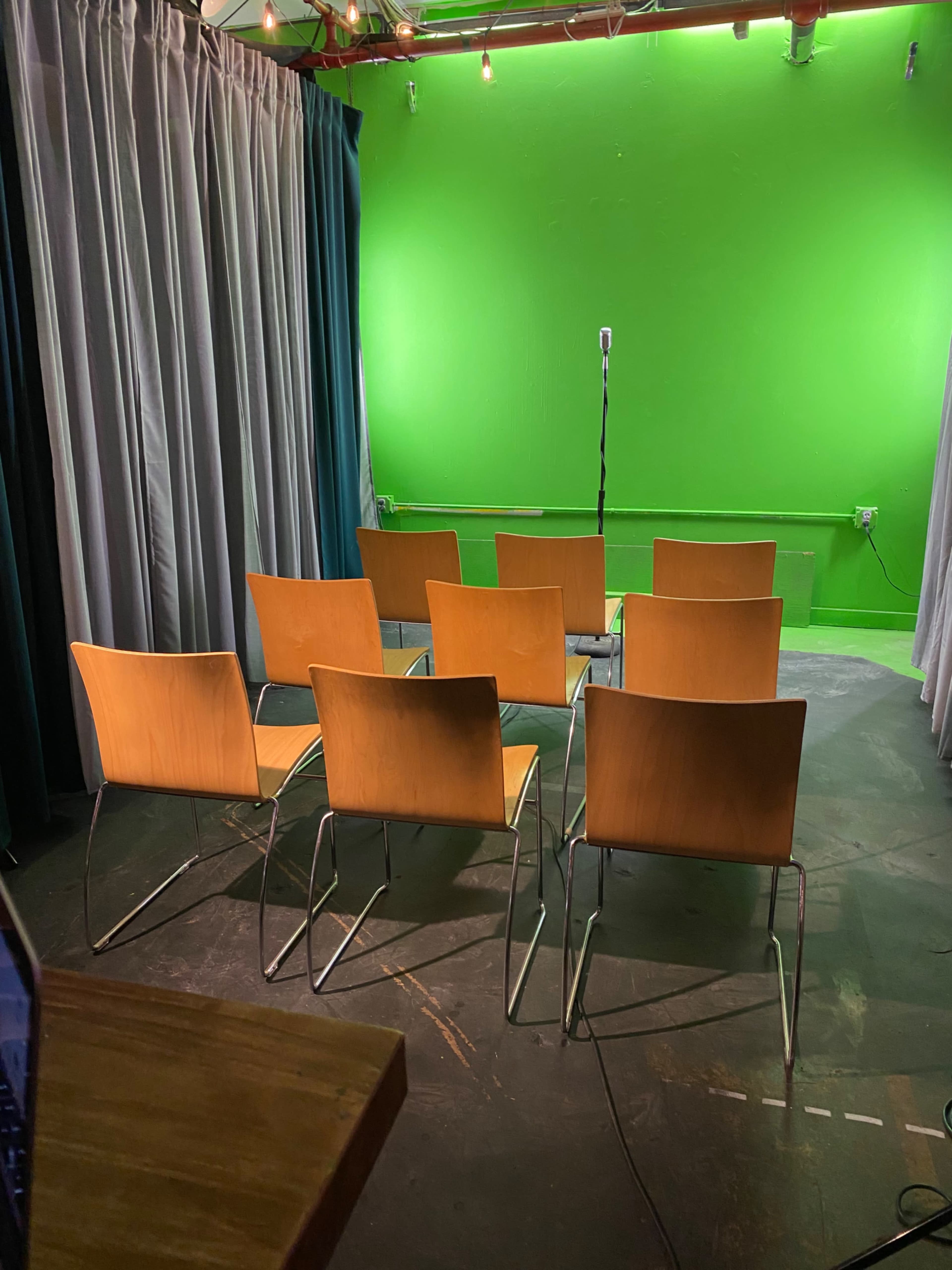 A set of eight wooden chairs arranged in front of a green backdrop with a microphone stand positioned in the center.