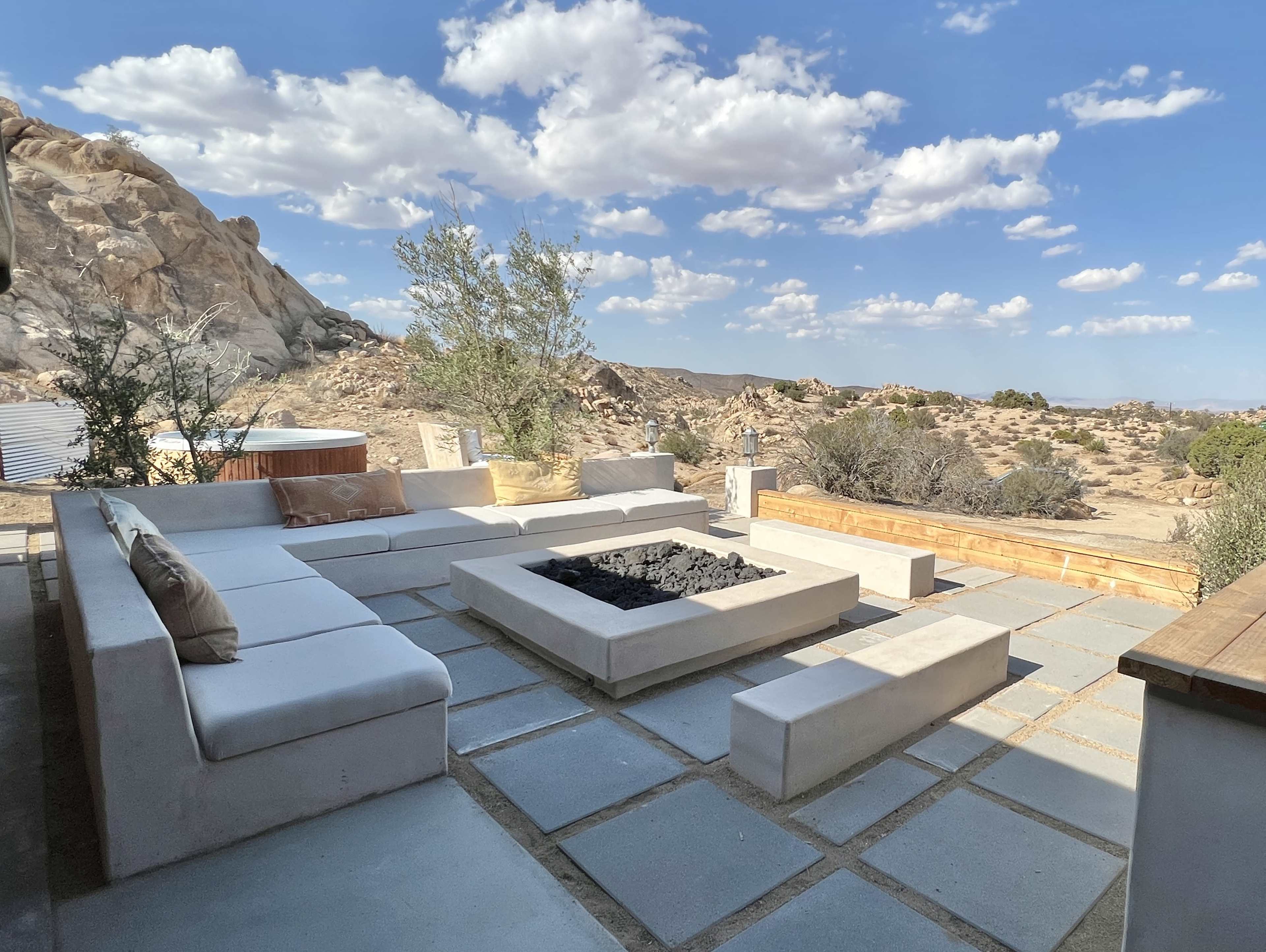 A modern outdoor seating area with a fire pit surrounded by stone slabs, set against a rocky desert landscape under a partly cloudy sky.