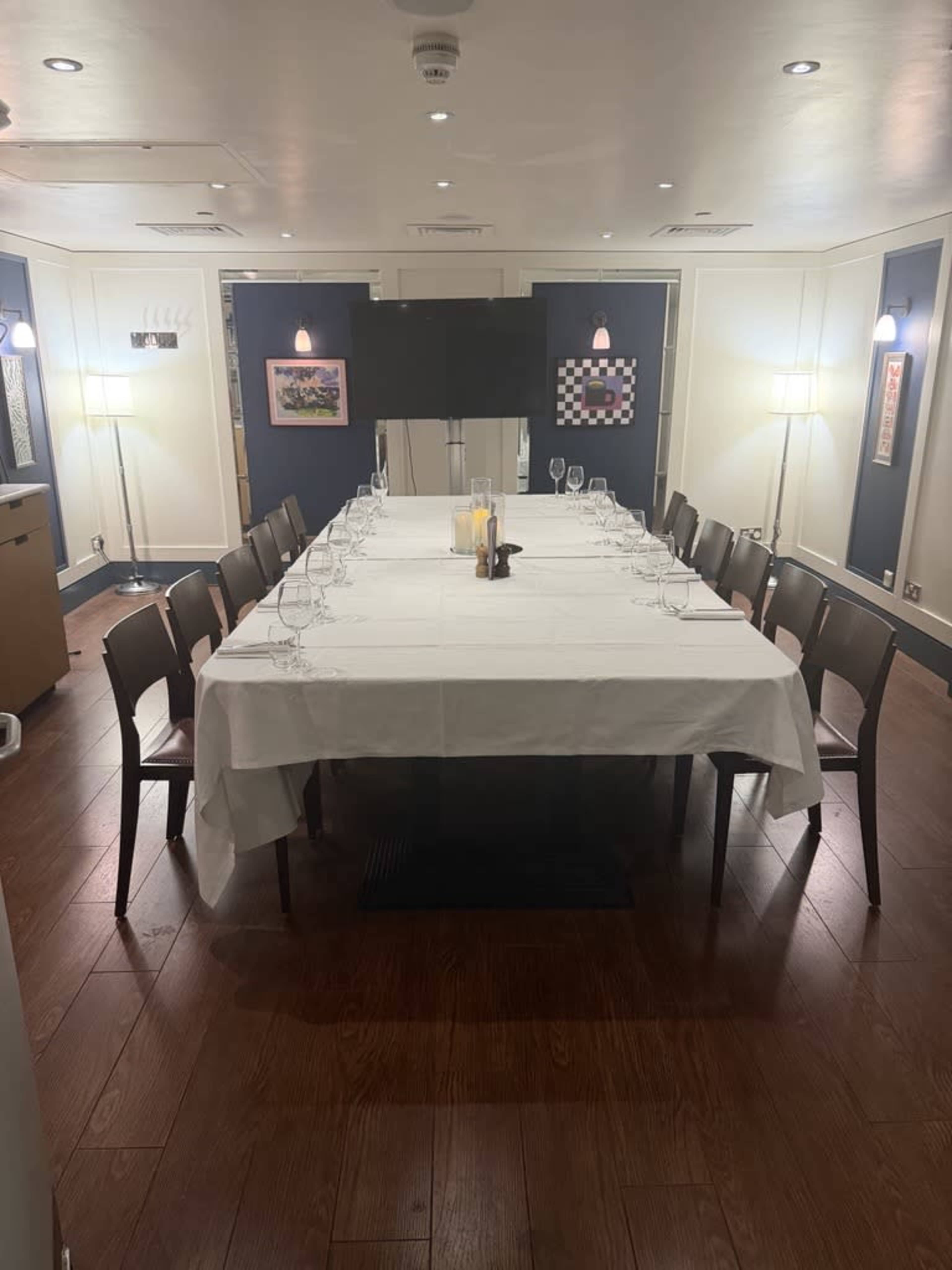 A long rectangular dining table is set with white tablecloth and glassware in a well-lit private dining room.