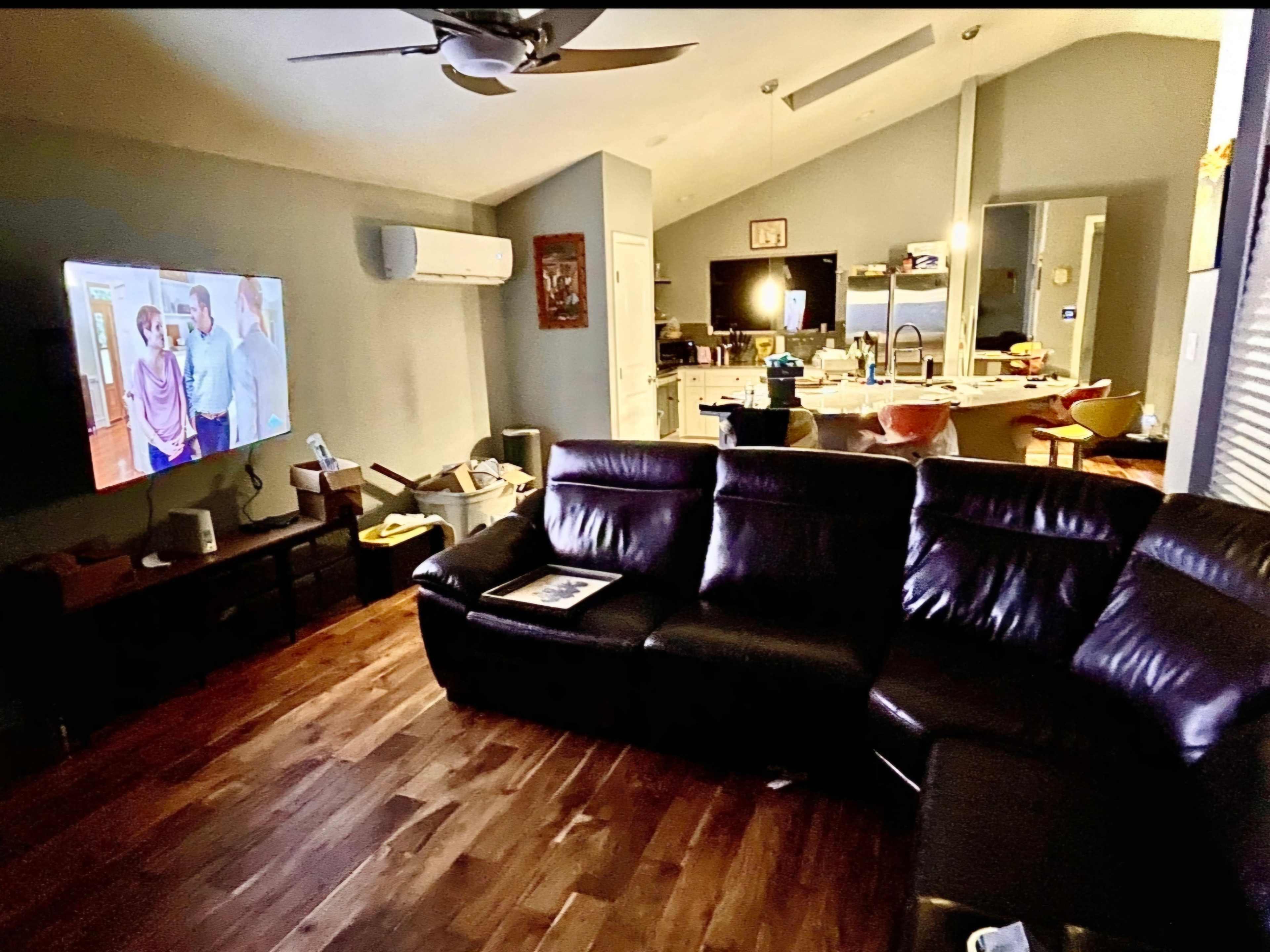 A spacious living room features a large sectional couch, a television displaying a scene, and a view of a kitchen area in the background.