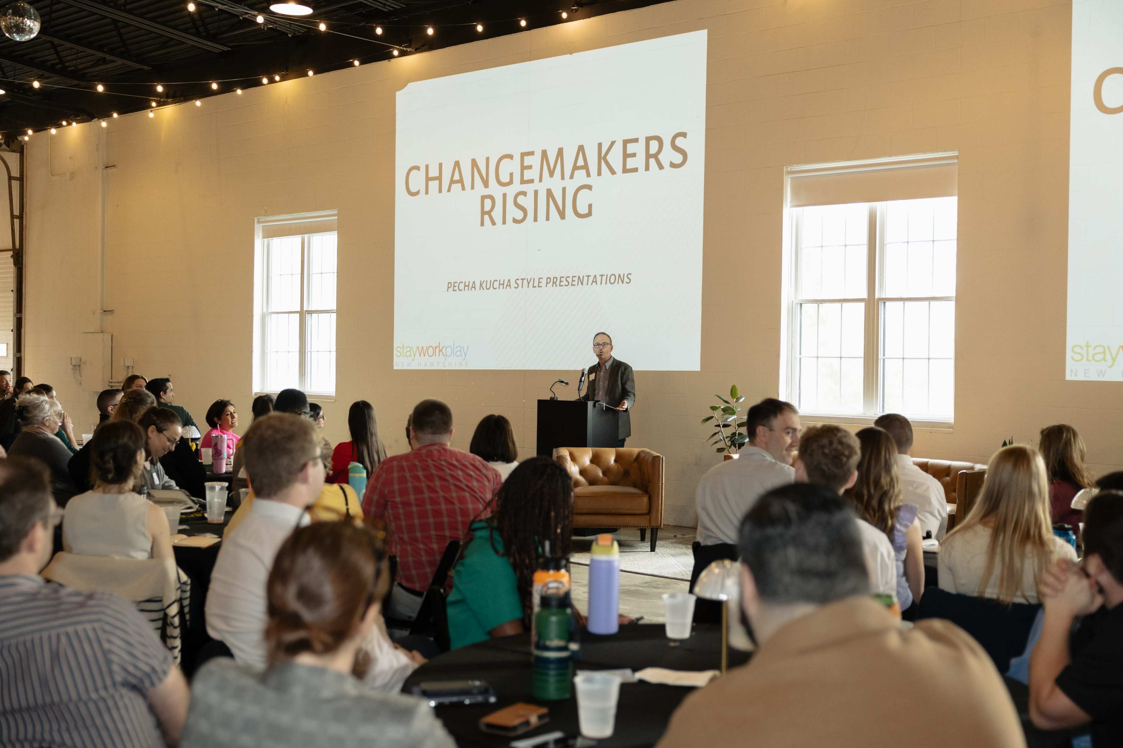 A speaker addresses an audience at a presentation event titled "Changemakers Rising" in a well-lit venue with large windows.