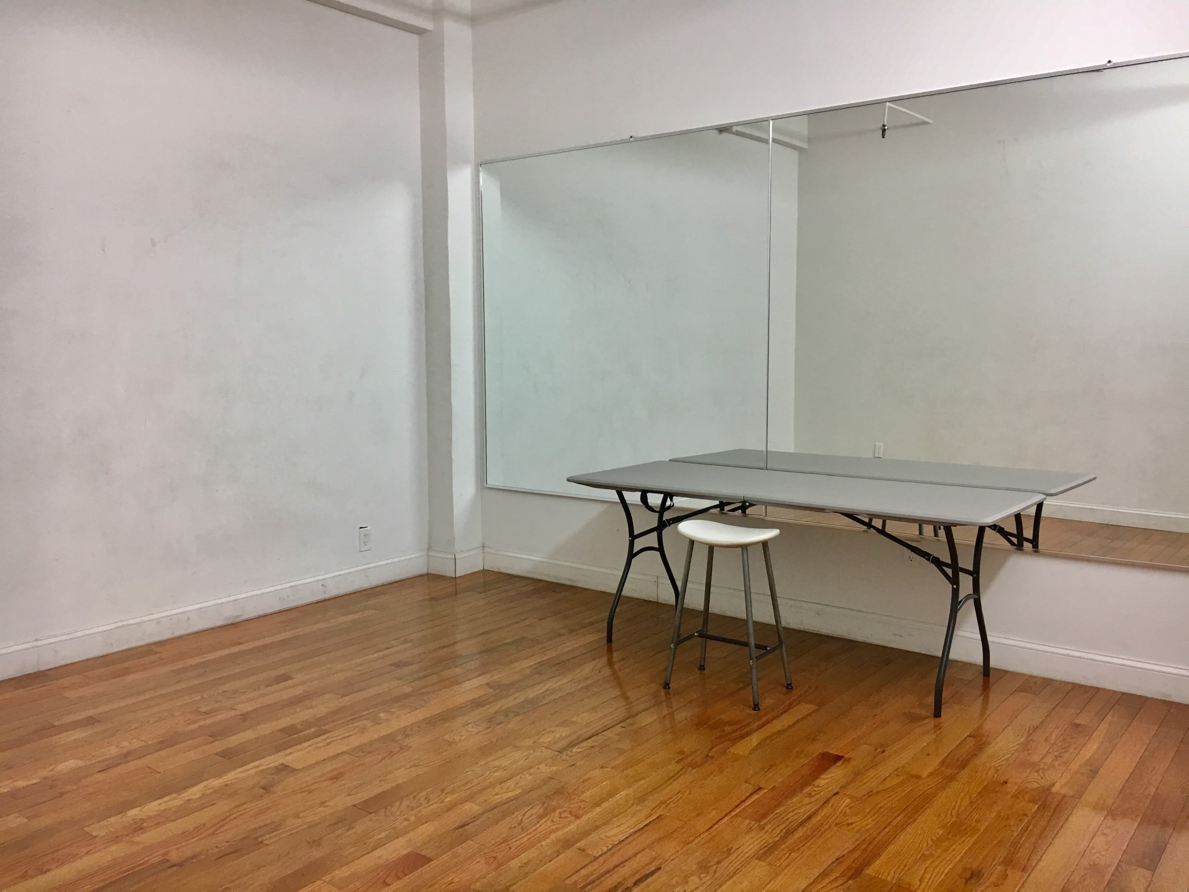 A simple empty room features a table and a stool facing a large mirror against a wall.