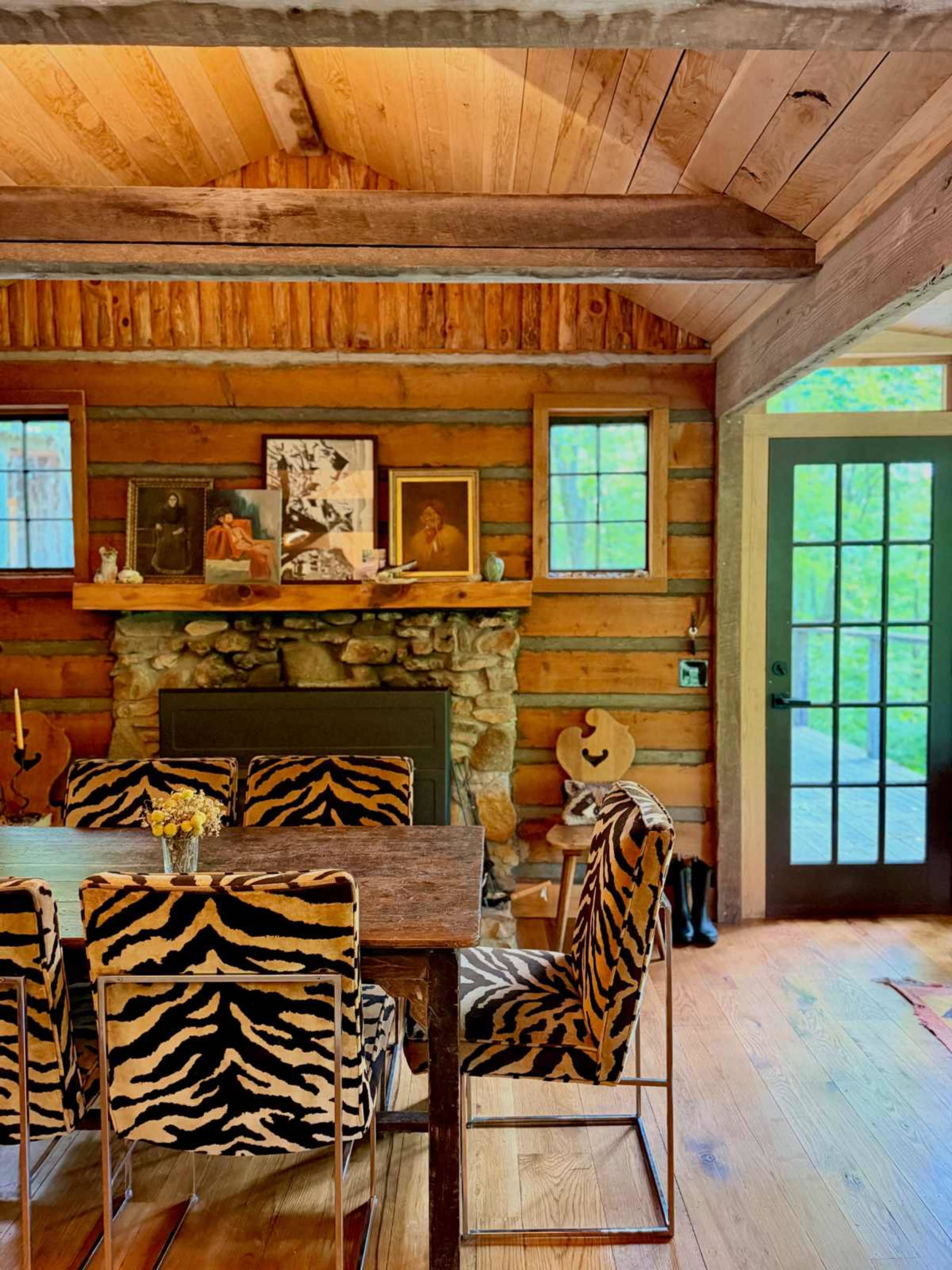 The image shows a rustic dining area with a wooden table surrounded by chairs upholstered in tiger-striped fabric, against a backdrop of log walls and framed artwork.