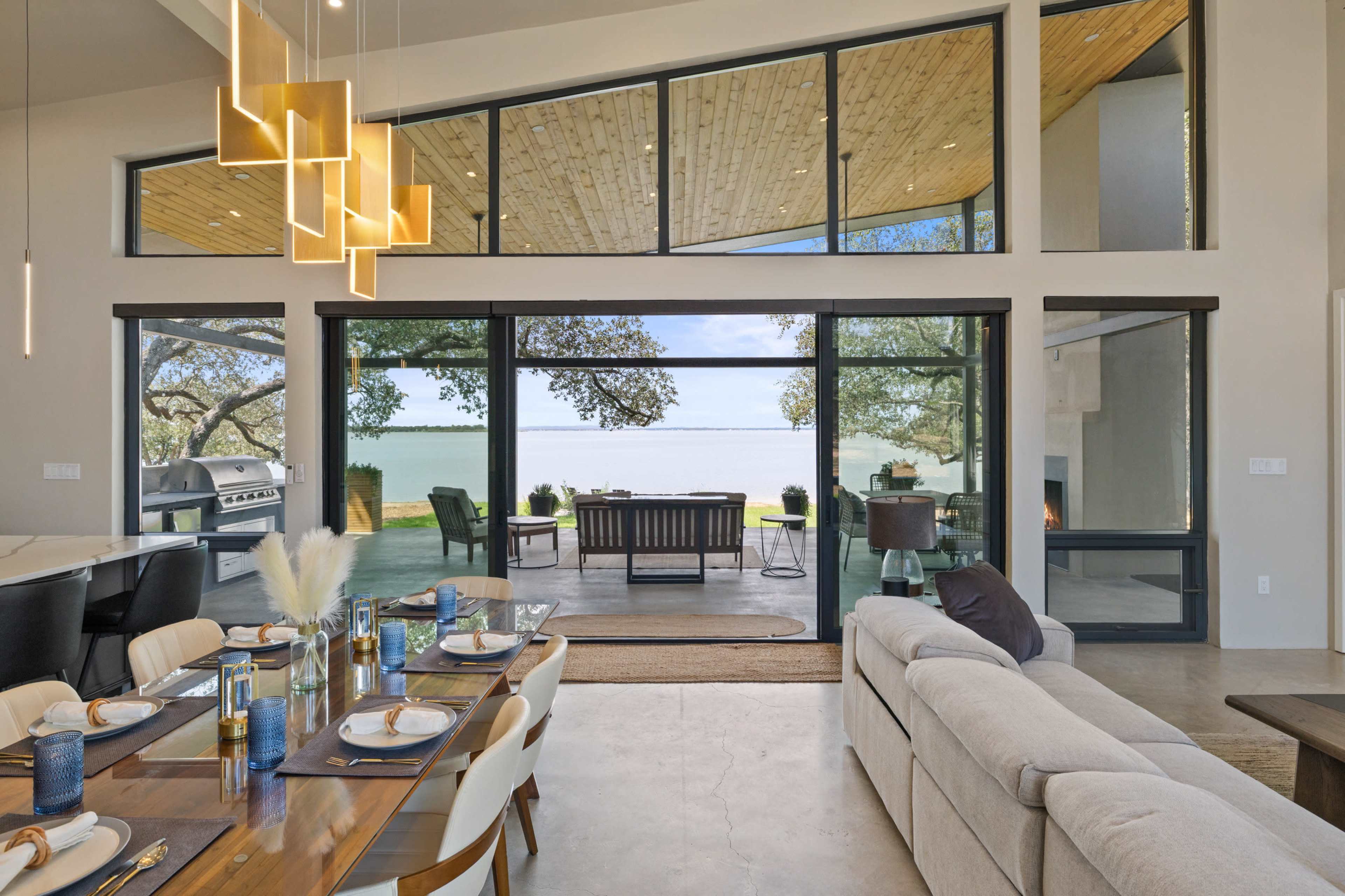 A modern dining area with a large wooden table overlooks a spacious outdoor patio and a waterfront view through floor-to-ceiling glass walls.