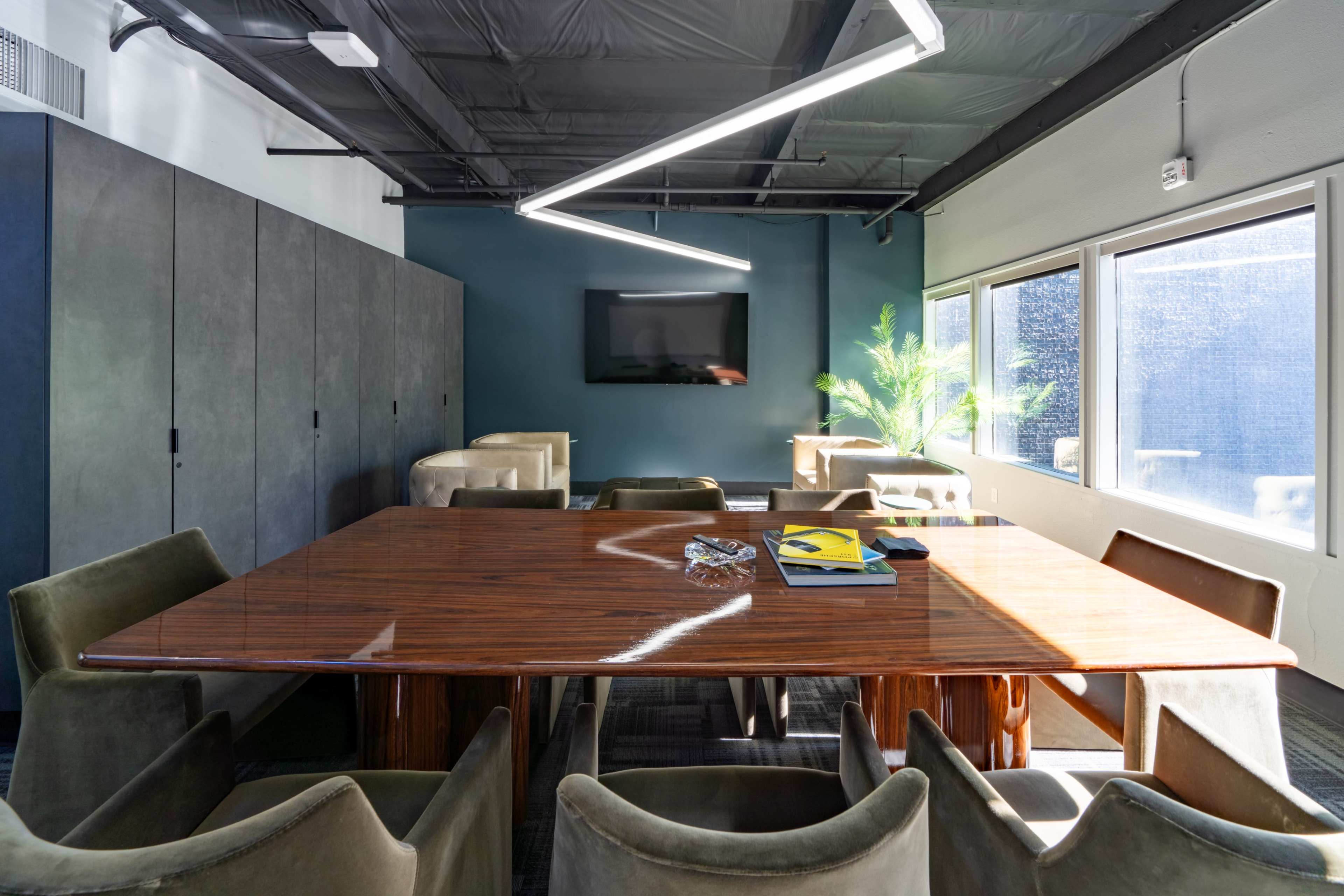 A modern conference room features a large wooden table surrounded by plush chairs, with a television mounted on the wall and natural light streaming through the windows.