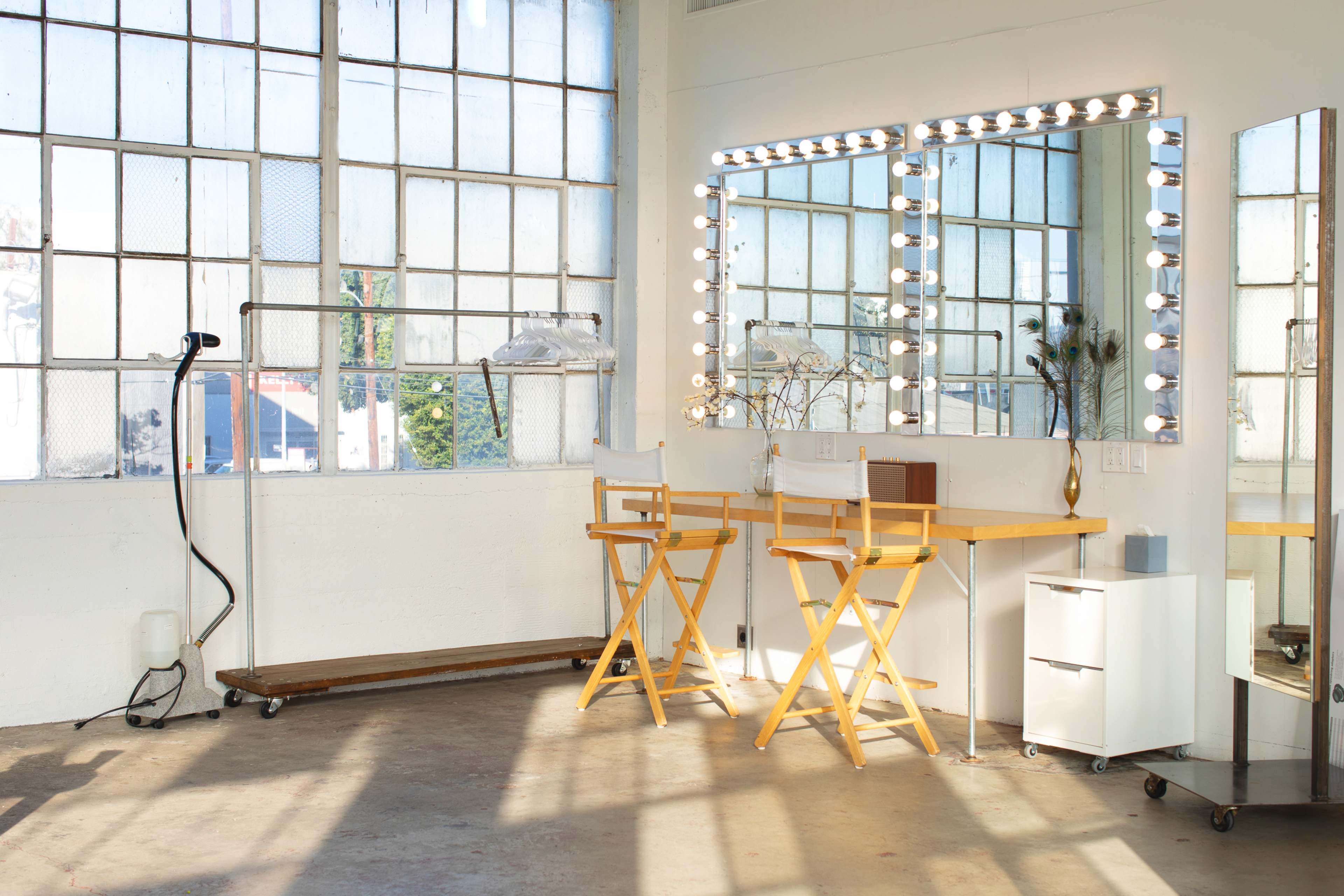 A bright, minimalist makeup room featuring two wooden director's chairs, a mirror with bulbs, and a white cabinet against a wall of large windows.