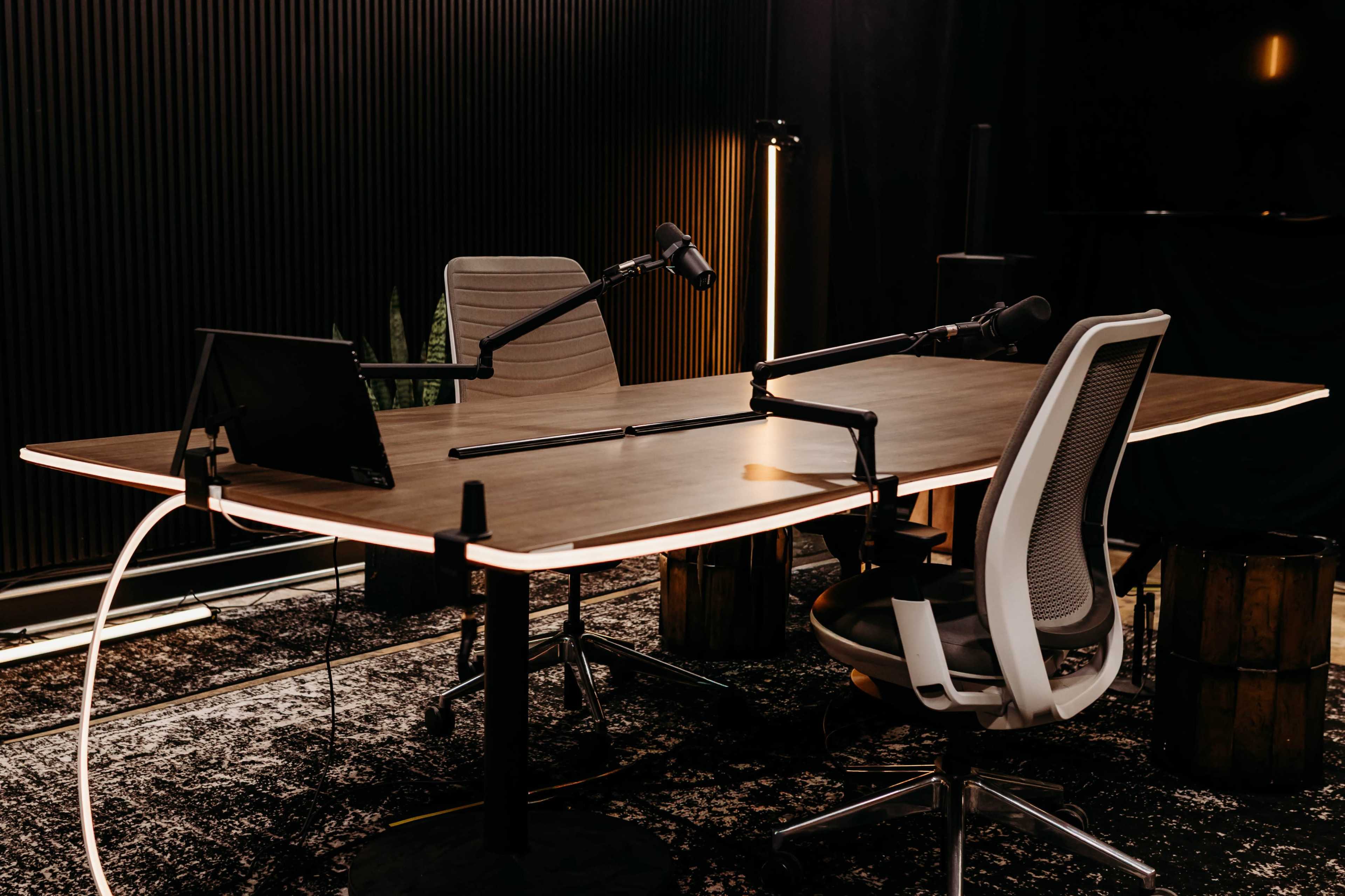 The image shows a modern podcast studio setup with a large wooden table, two microphones on adjustable arms, and a comfortable office chair, all arranged in a dimly lit environment.