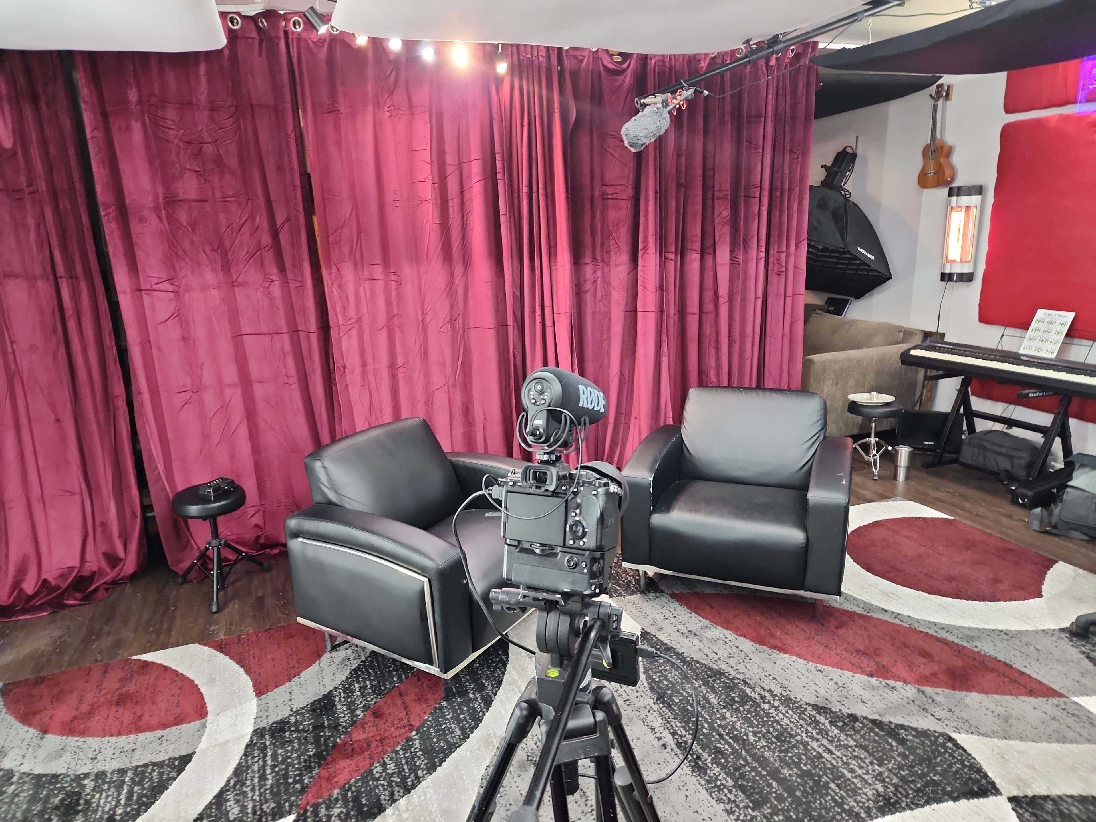 A recording setup with two black chairs in front of a red curtain, a microphone overhead, and a camera on a tripod.