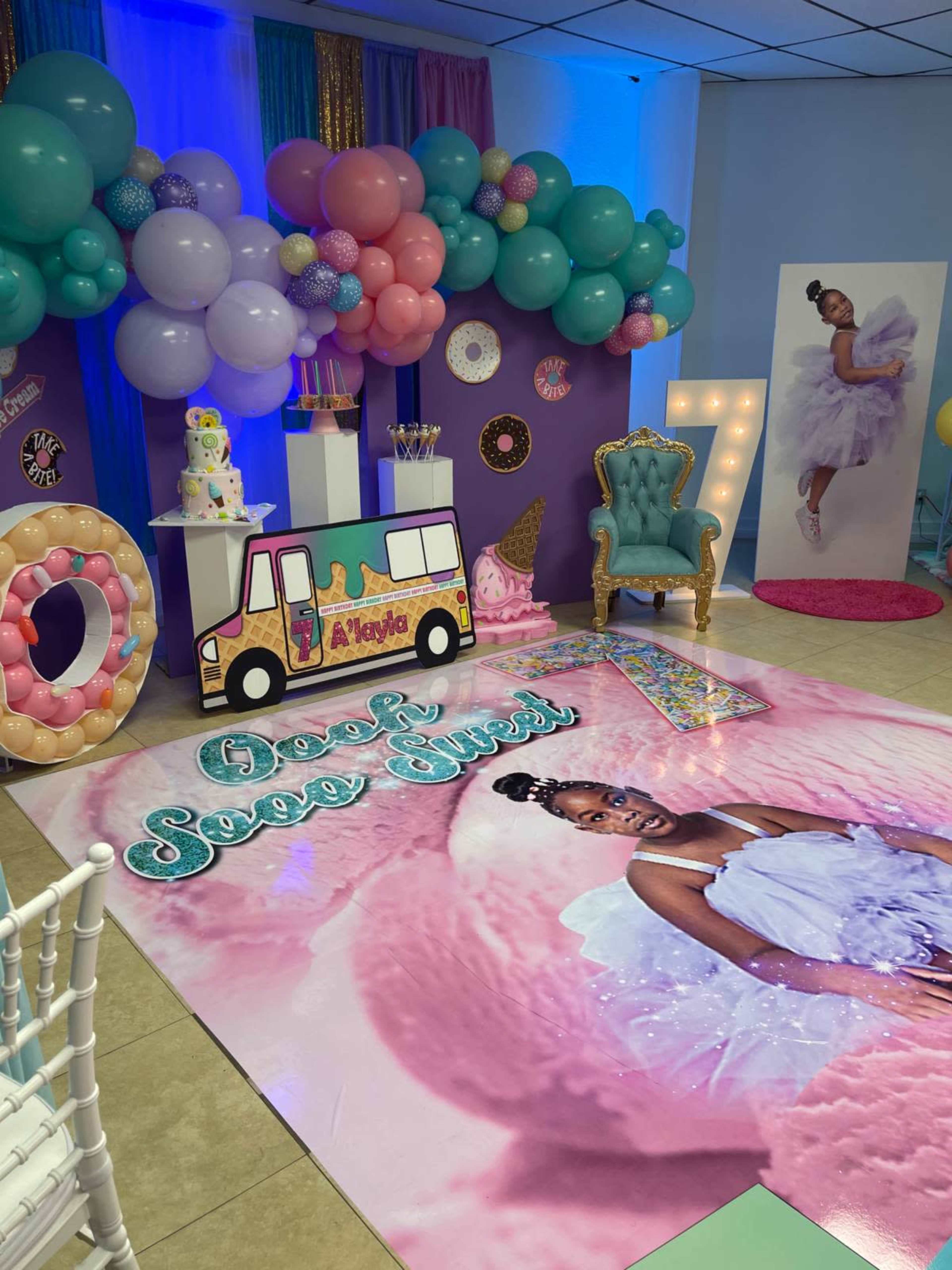 The scene features a colorful party setup with balloons, a decorated dessert table, and a large floor display featuring a portrait of a girl in a white dress.