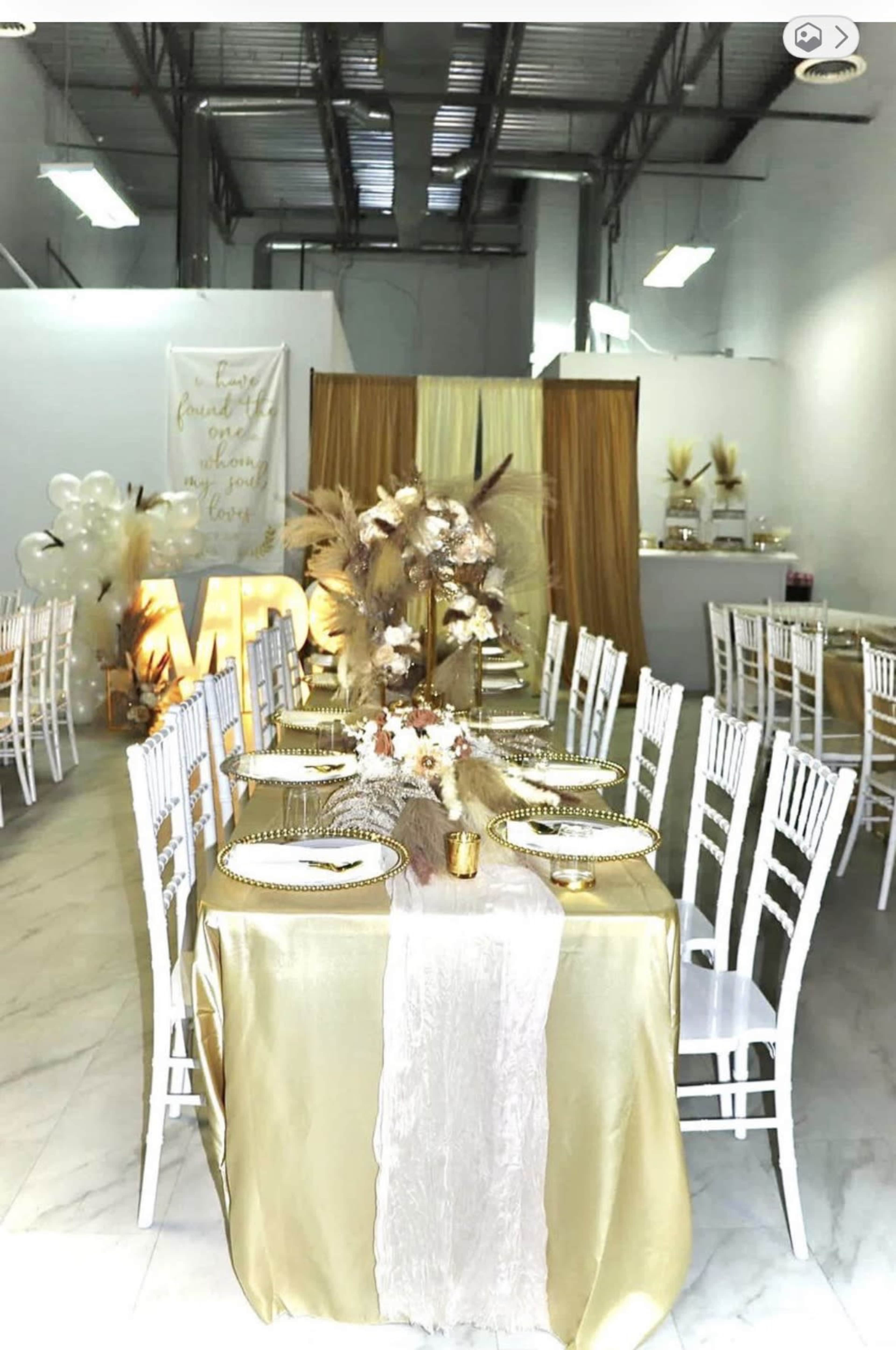 A banquet hall is set up with a gold-themed table arrangement, featuring white chairs and decorations that include dried plants and balloons.