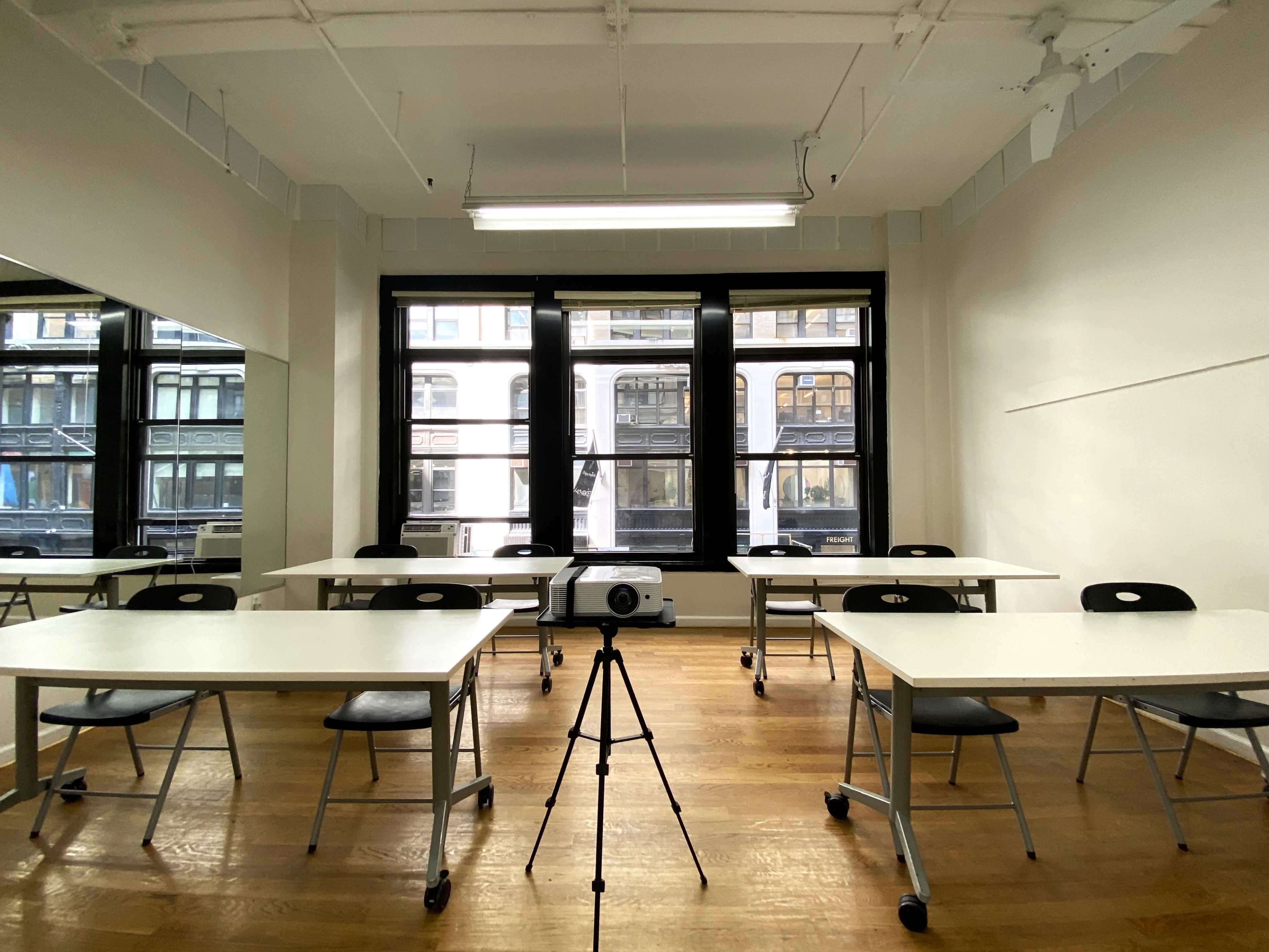 A bright, empty classroom is set up with several tables and chairs facing a projector on a tripod, with large windows providing a view of the street outside.