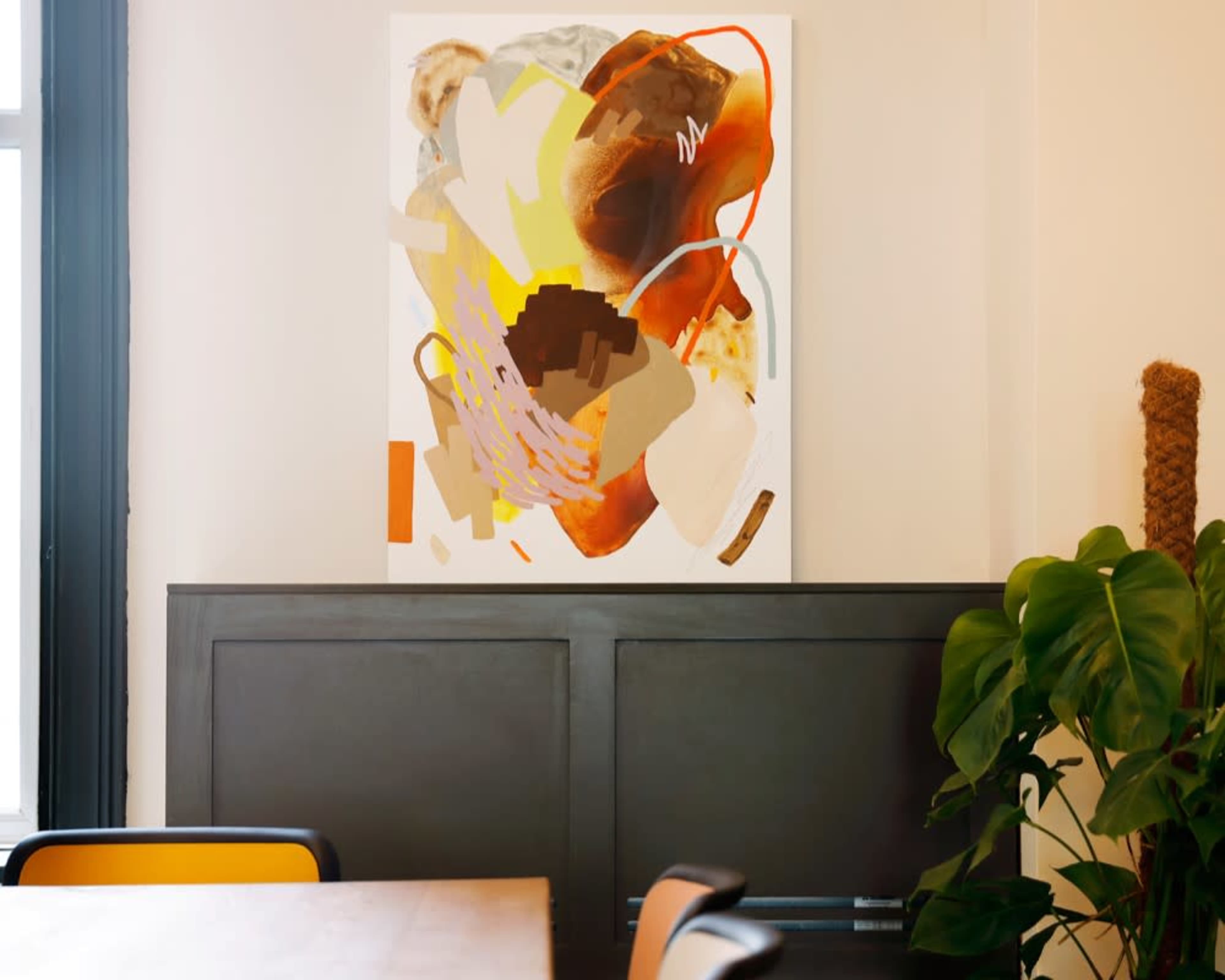 A large abstract painting with vibrant colors hangs on a light-colored wall beside a dark panel, while a potted plant sits nearby in a minimalist dining area.