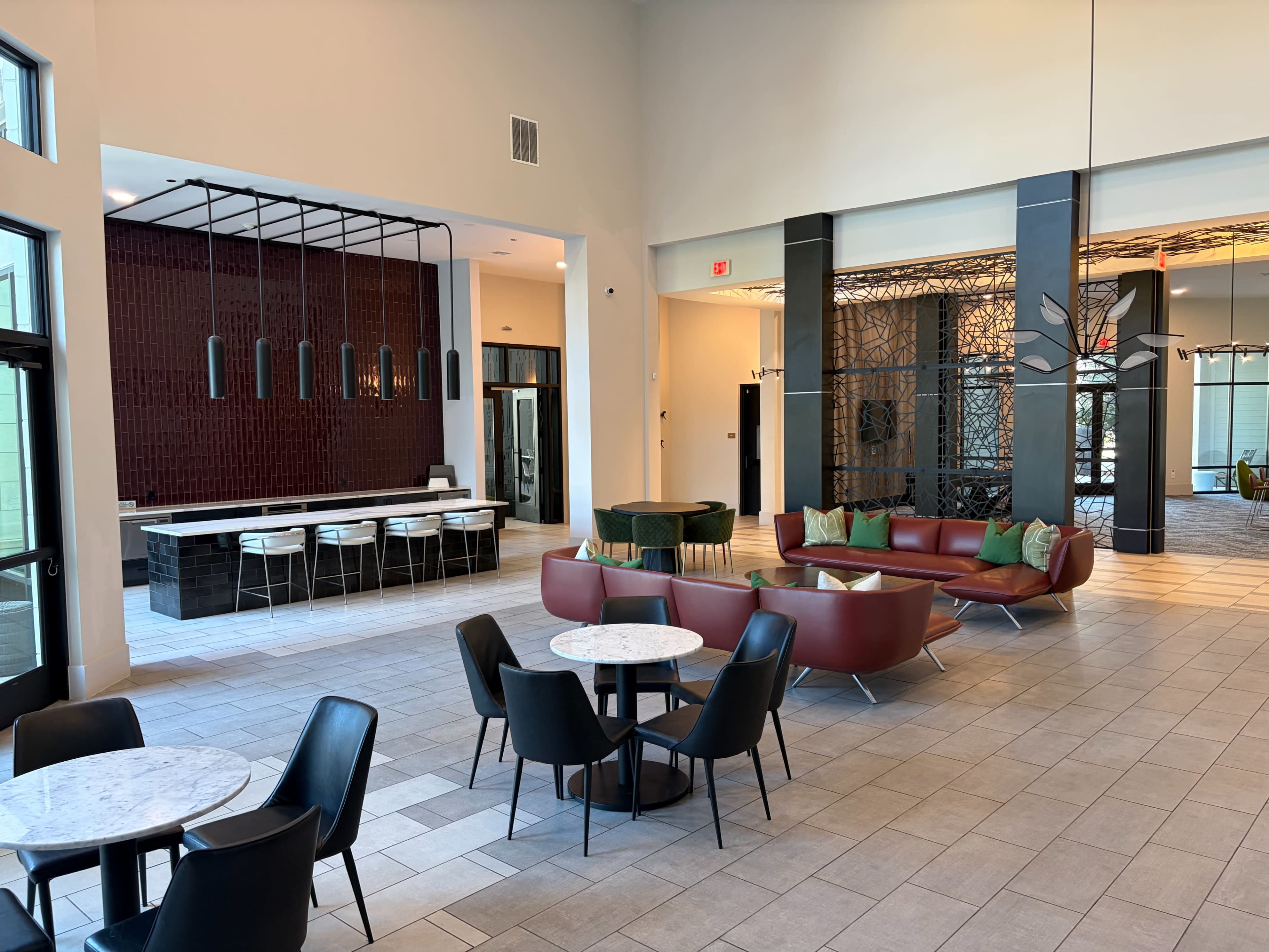 The image shows a modern lobby area featuring red and green seating arrangements, marble-top tables, and a bar with pendant lights in the background.