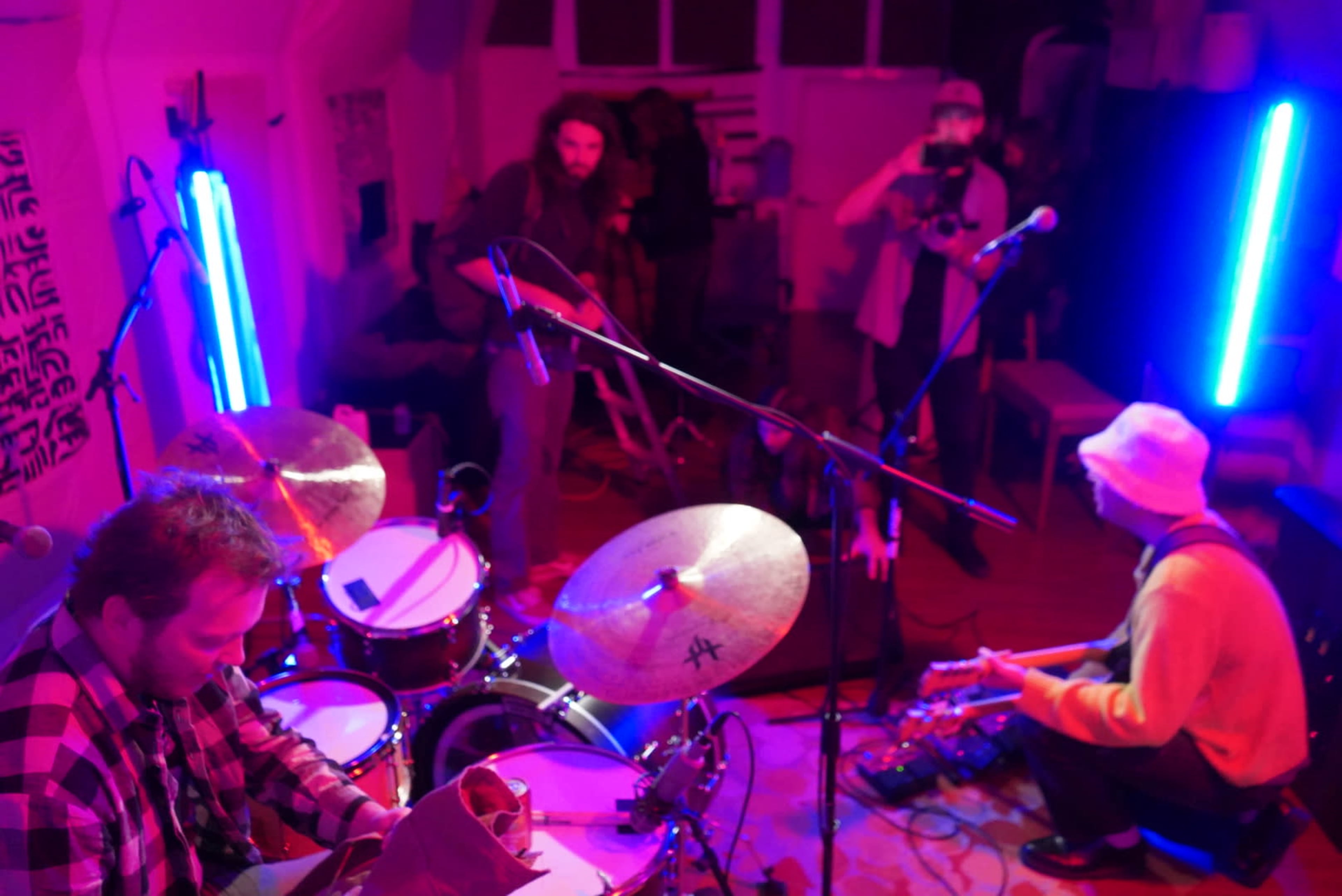 A drummer and a guitarist are performing in a small, brightly lit room with colorful LED lights and a few people capturing the moment on camera.