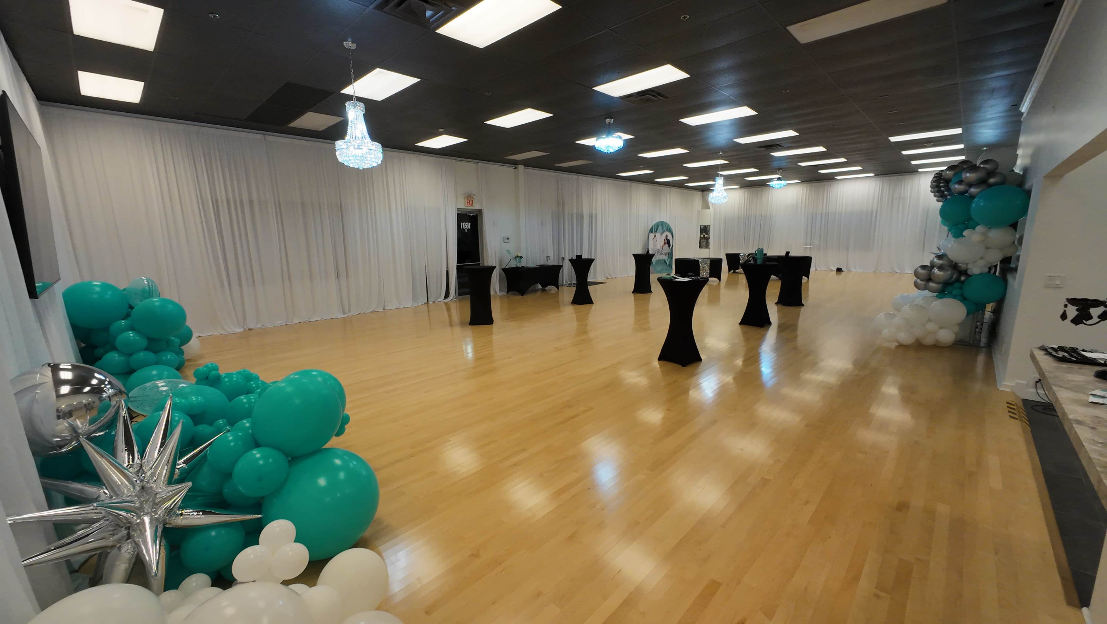 The spacious event hall features black cocktail tables, decorative balloons in teal and white, and elegant lighting fixtures.
