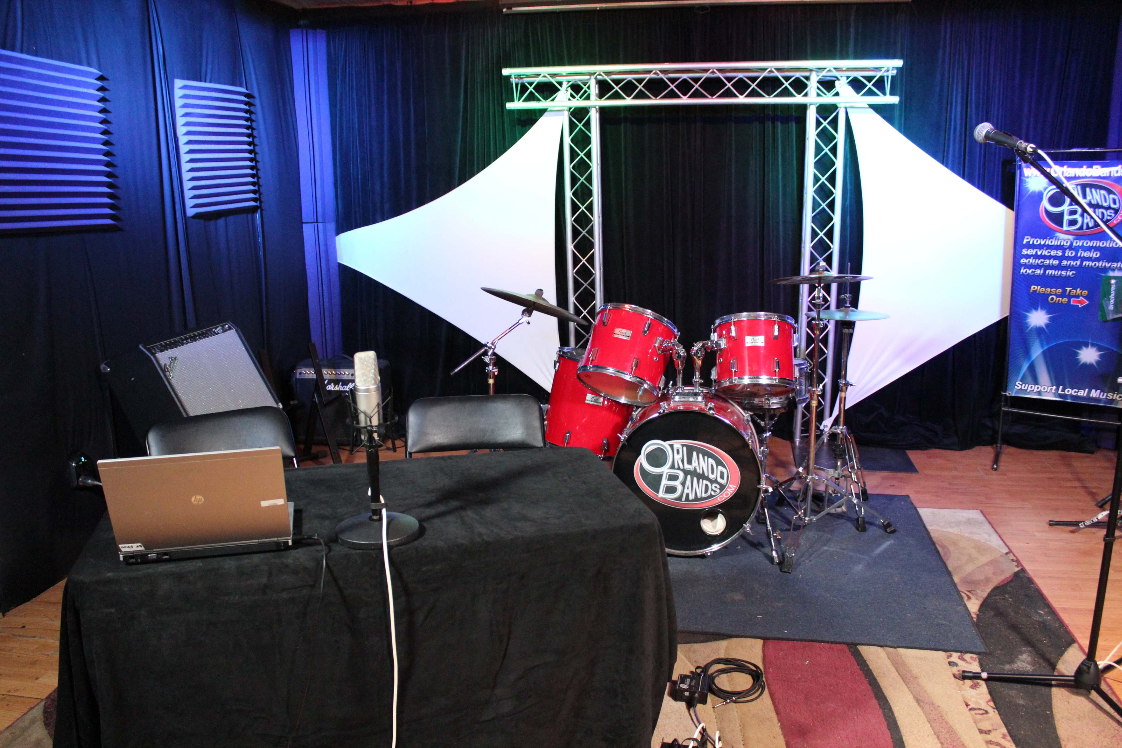 The image shows a small stage setup with a drum kit, microphones, and a laptop on a table, surrounded by black curtains and lighting.