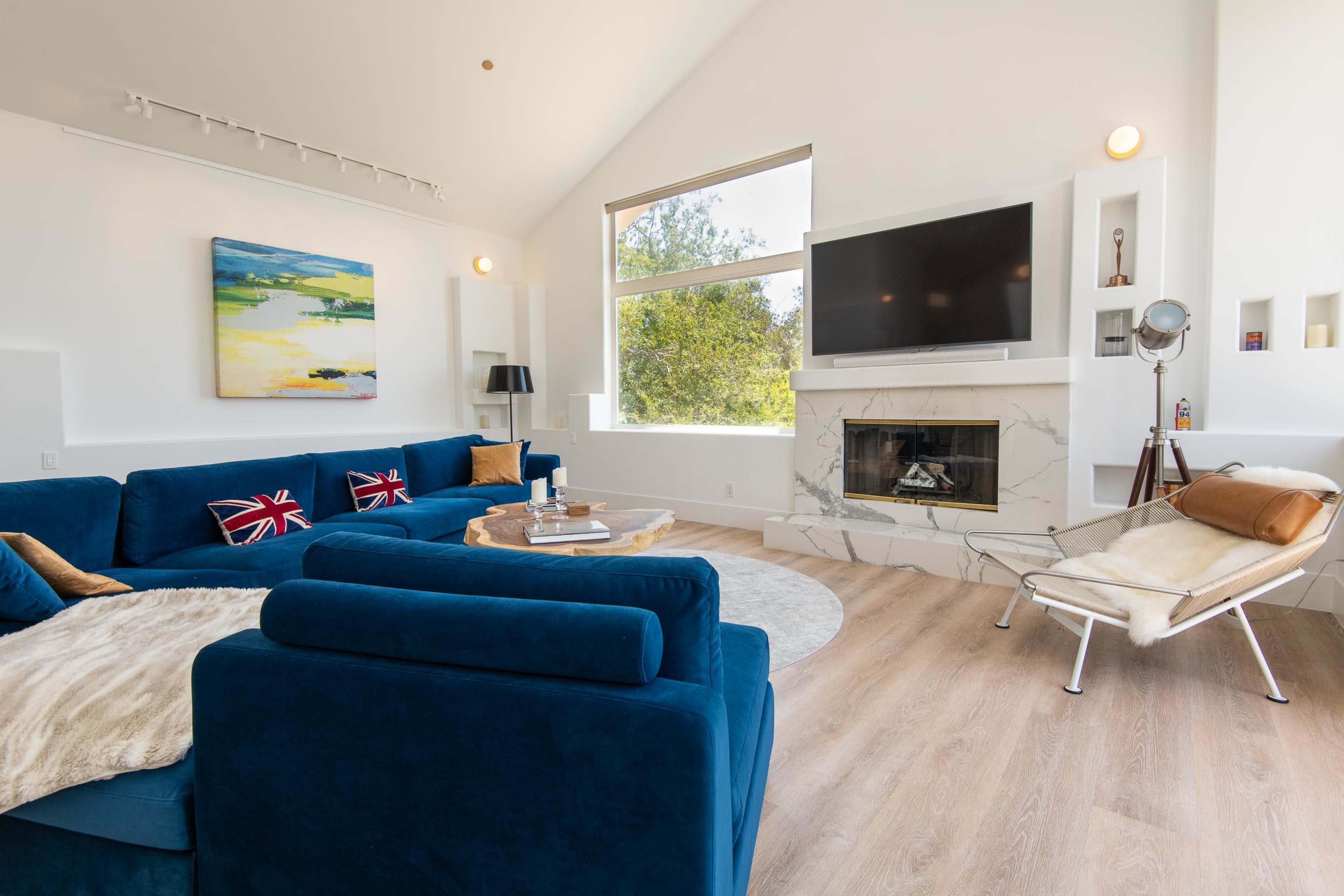 The living room features a blue sectional sofa, a marble fireplace, a large television mounted on the wall, and a comfortable chair beside a window with natural light.