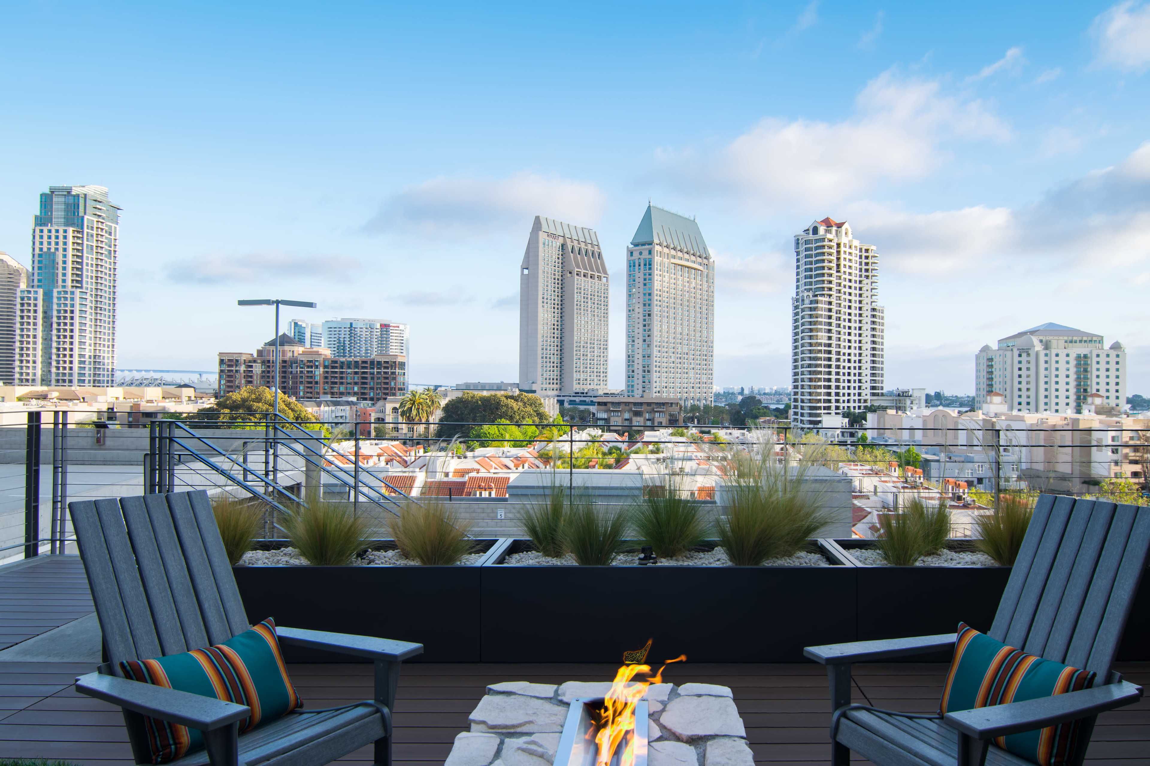 A rooftop terrace features two wooden chairs facing a fire pit, with a skyline of high-rise buildings in the background.