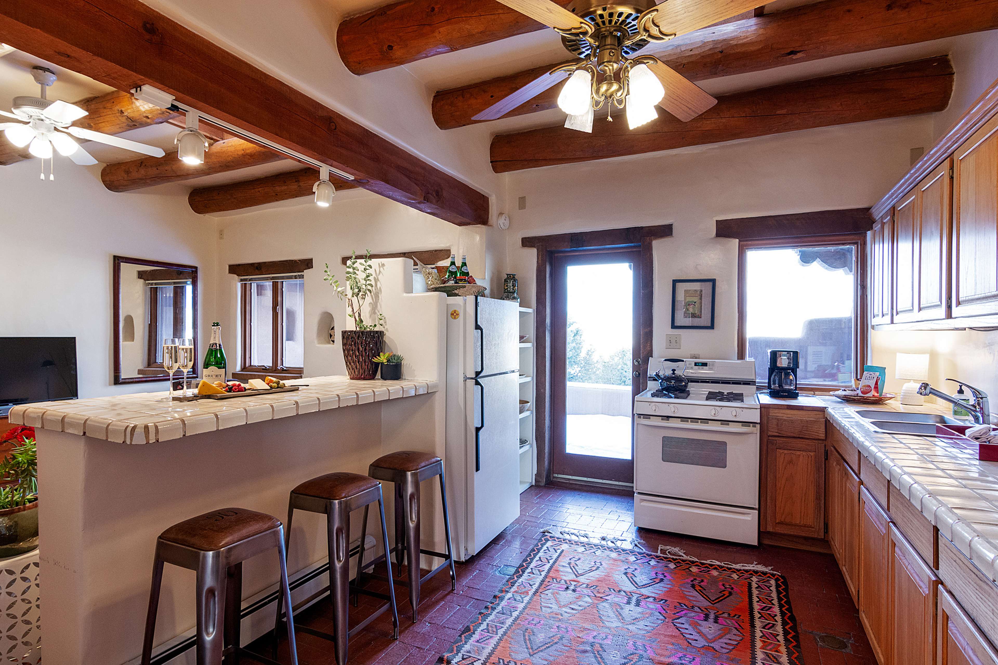A rustic kitchen features wooden beams, a white tile countertop with bar stools, and a view of a patio through the doorway.