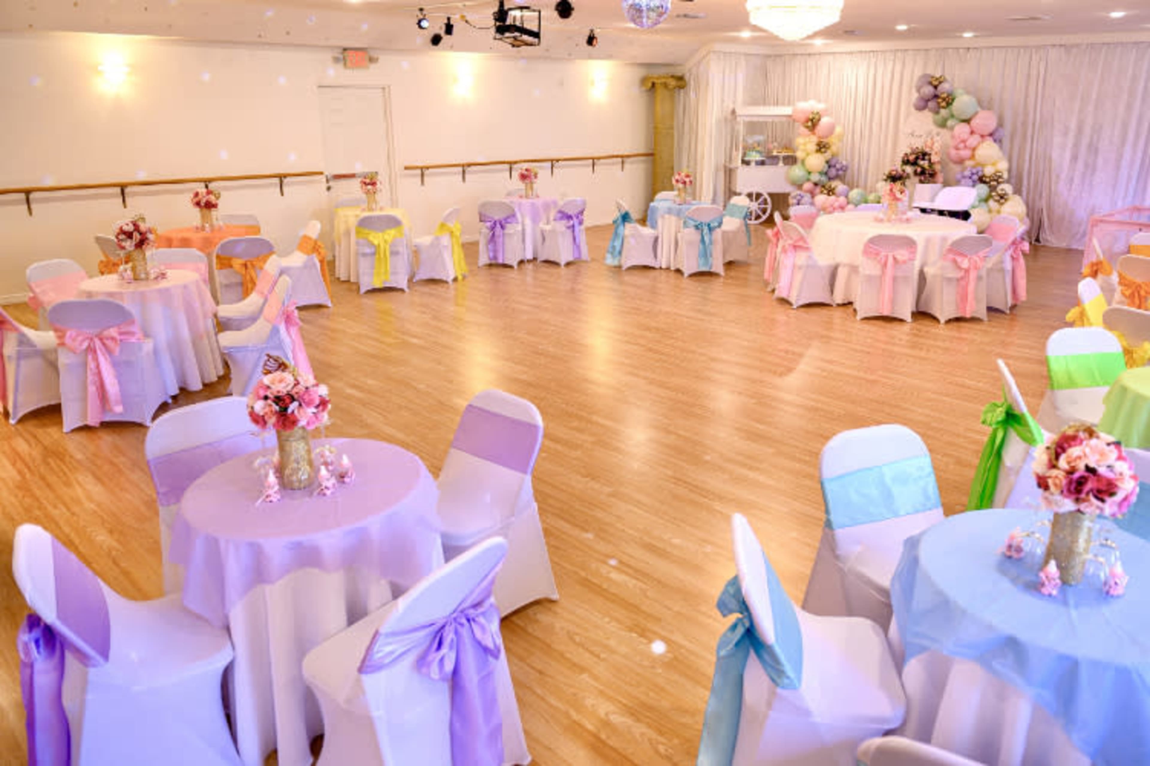 A decorated event space features round tables with colorful tablecloths and sashes, surrounded by chairs, and adorned with floral centerpieces.