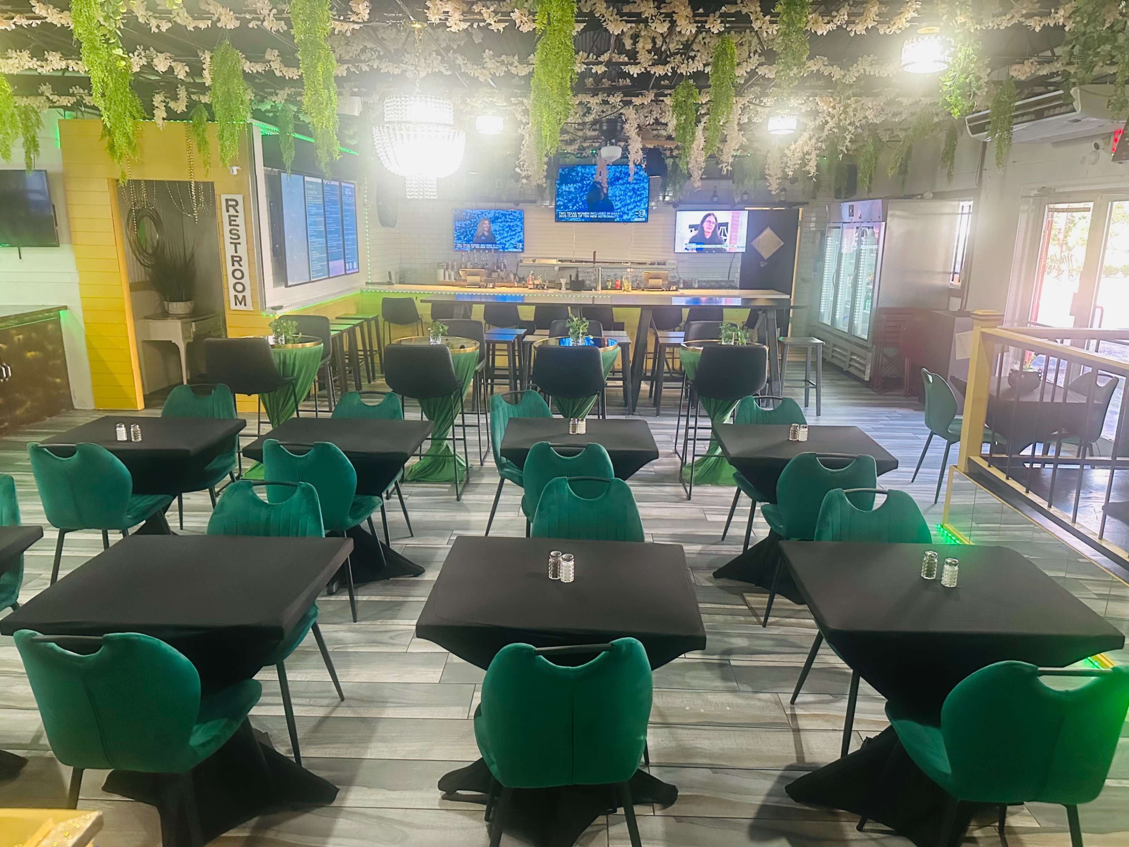 The image shows a modern restaurant setup with green chairs around black tables, a bar area in the background, and decorative hanging plants.