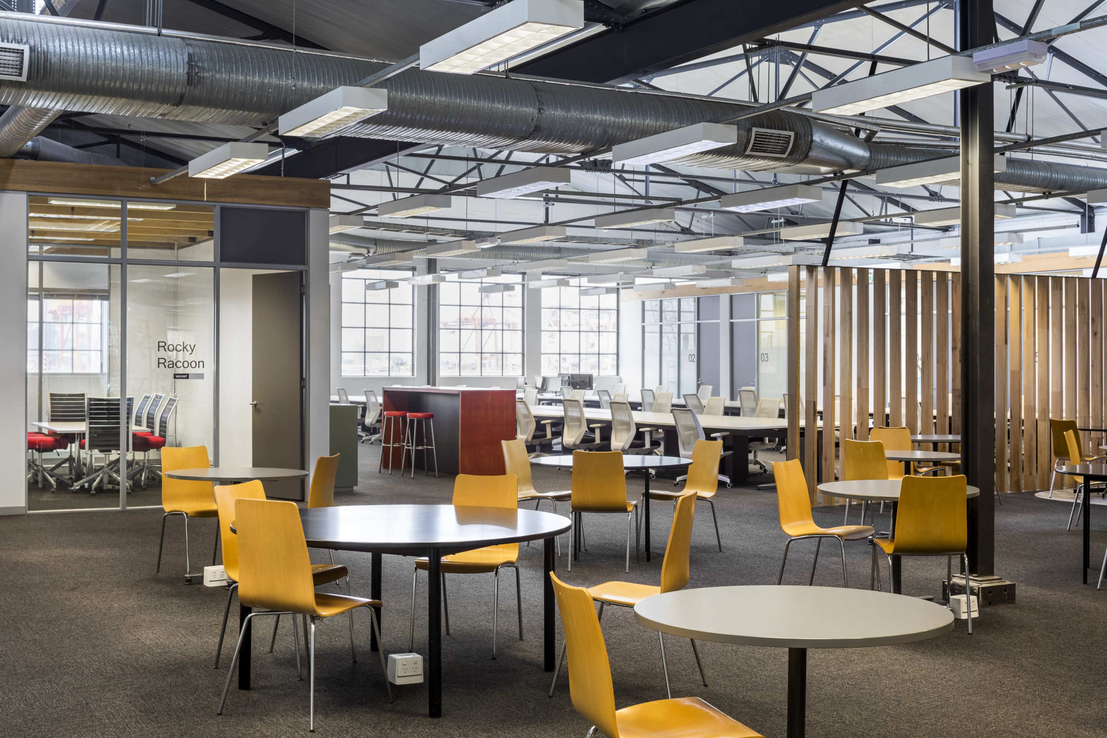 The image shows a modern office space with several round tables and chairs, large windows allowing natural light, and distinct areas for collaboration.